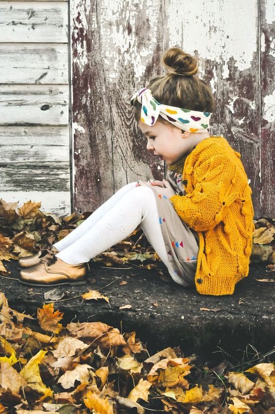 Child in mustard yellow sweater sits among autumn leaves wearing patterned headband.