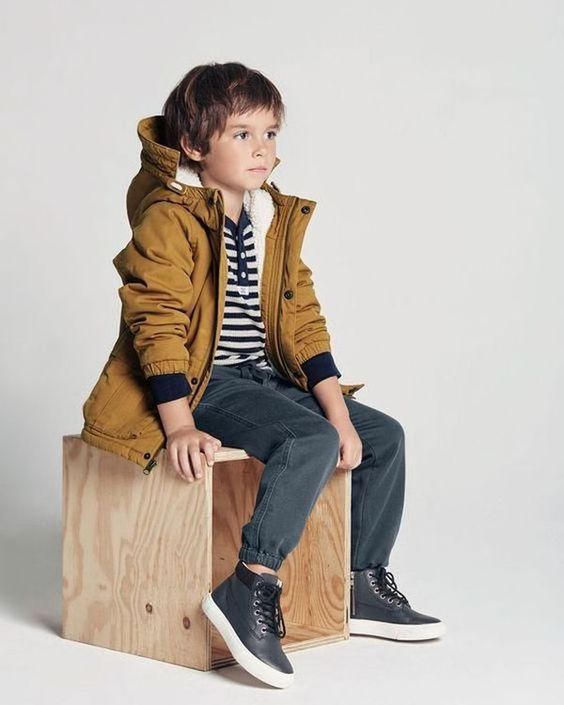 Child in brown hooded jacket and striped shirt sits on wooden crate against white background.