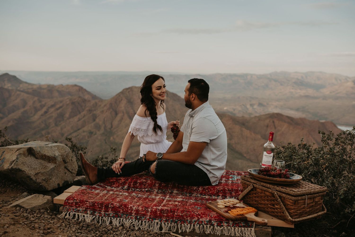Couple enjoying picnic on red blanket with desert mountain views.