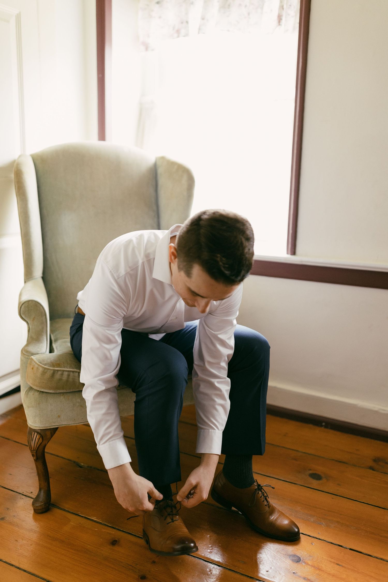 Someone adjusts formal dress shoes while sitting in an elegant room with hardwood floors.