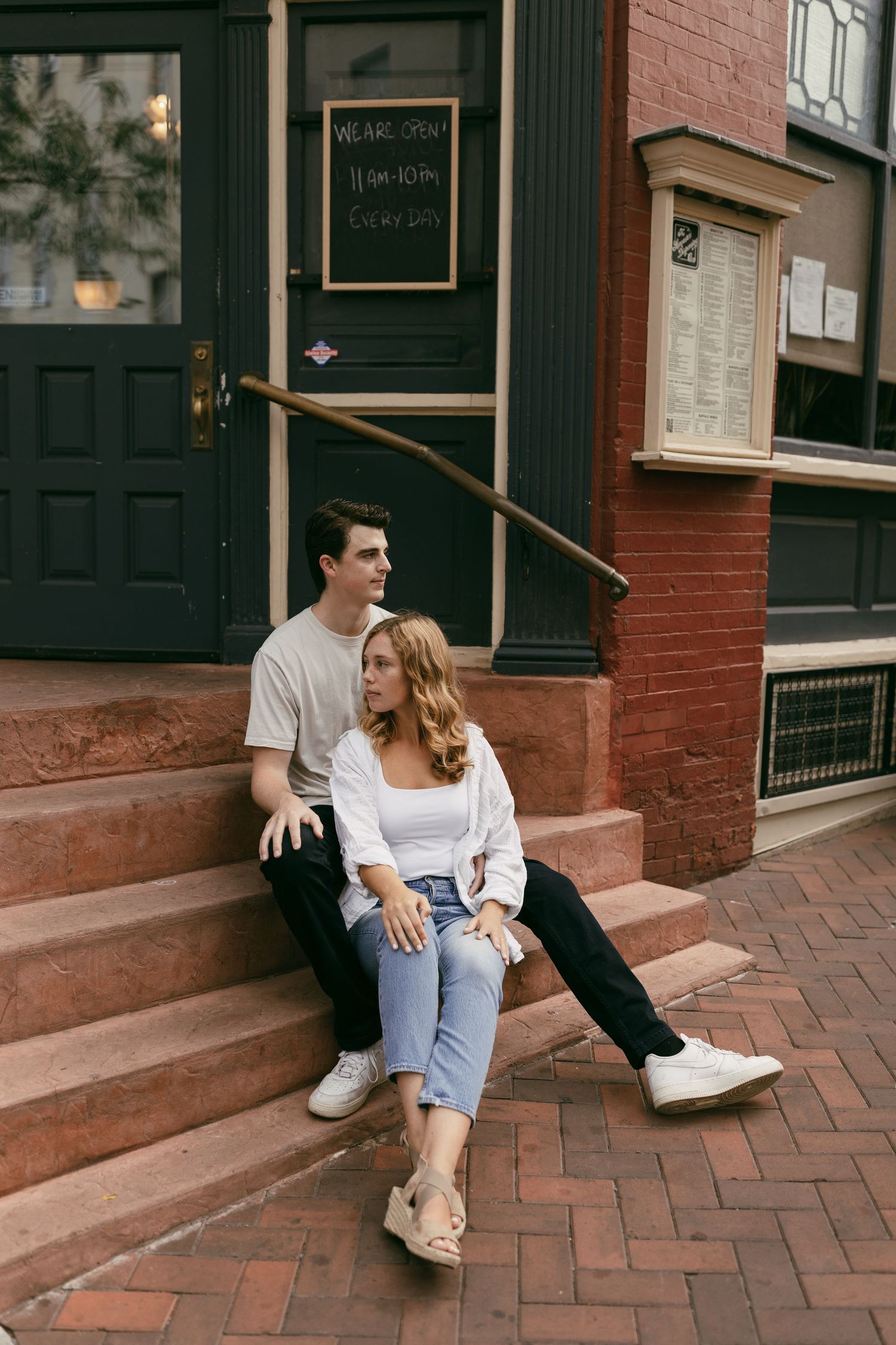 A casual couple sits on brick steps outside a charming storefront.