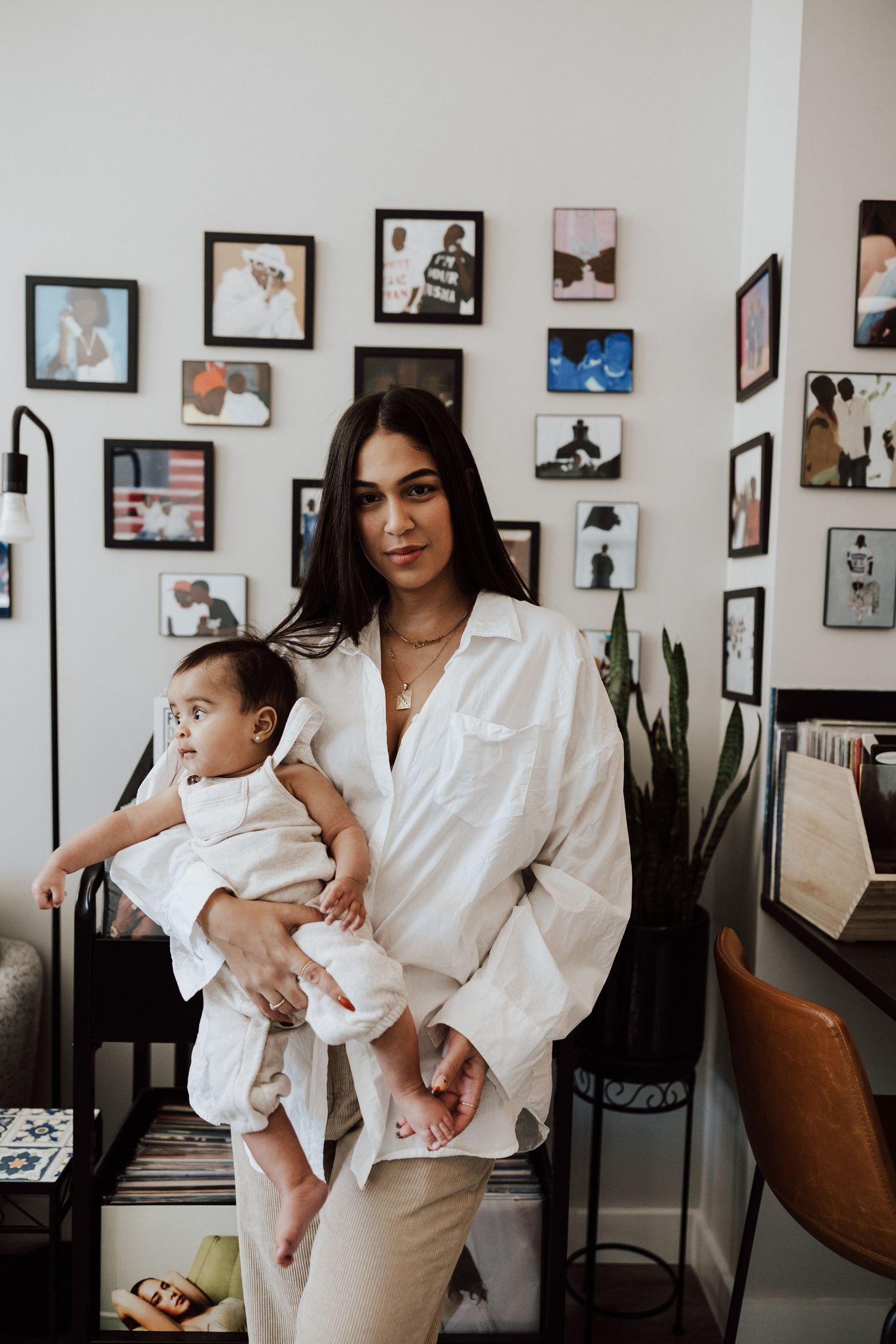 A woman in a white oversized shirt holds a baby in a stylish room with a gallery wall of framed photos behind her. Miranda Photo & Co