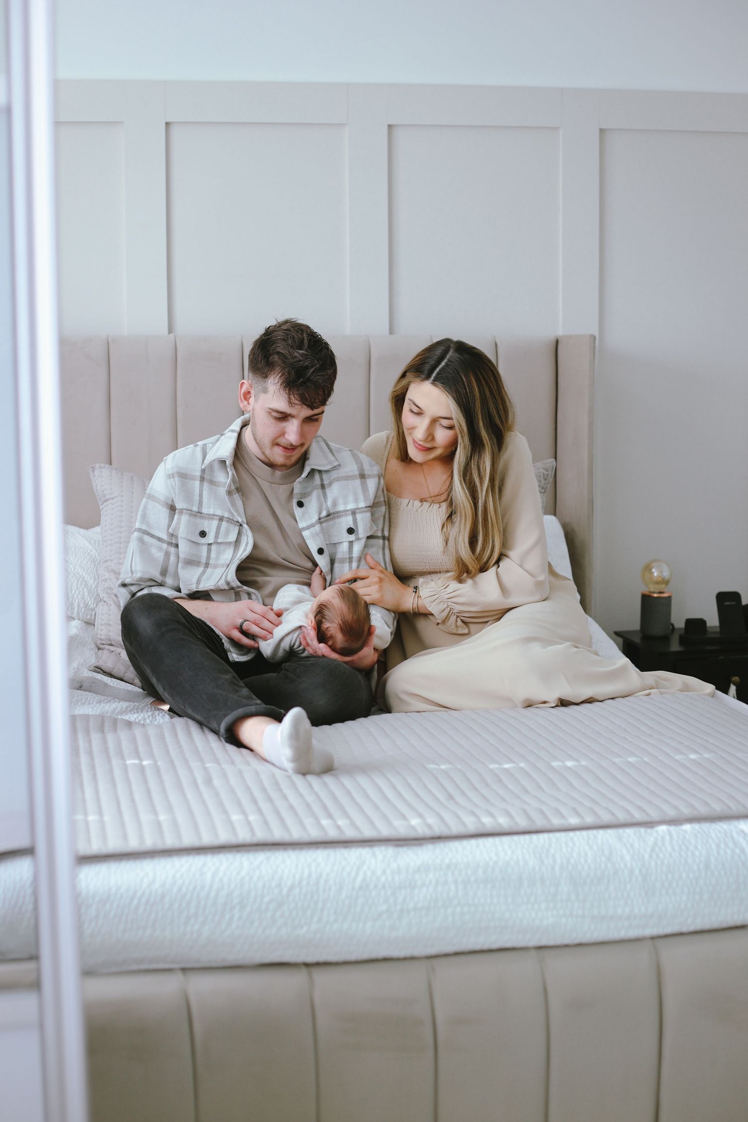 A couple sits on a white bed with their newborn baby in a cozy bedroom setting.