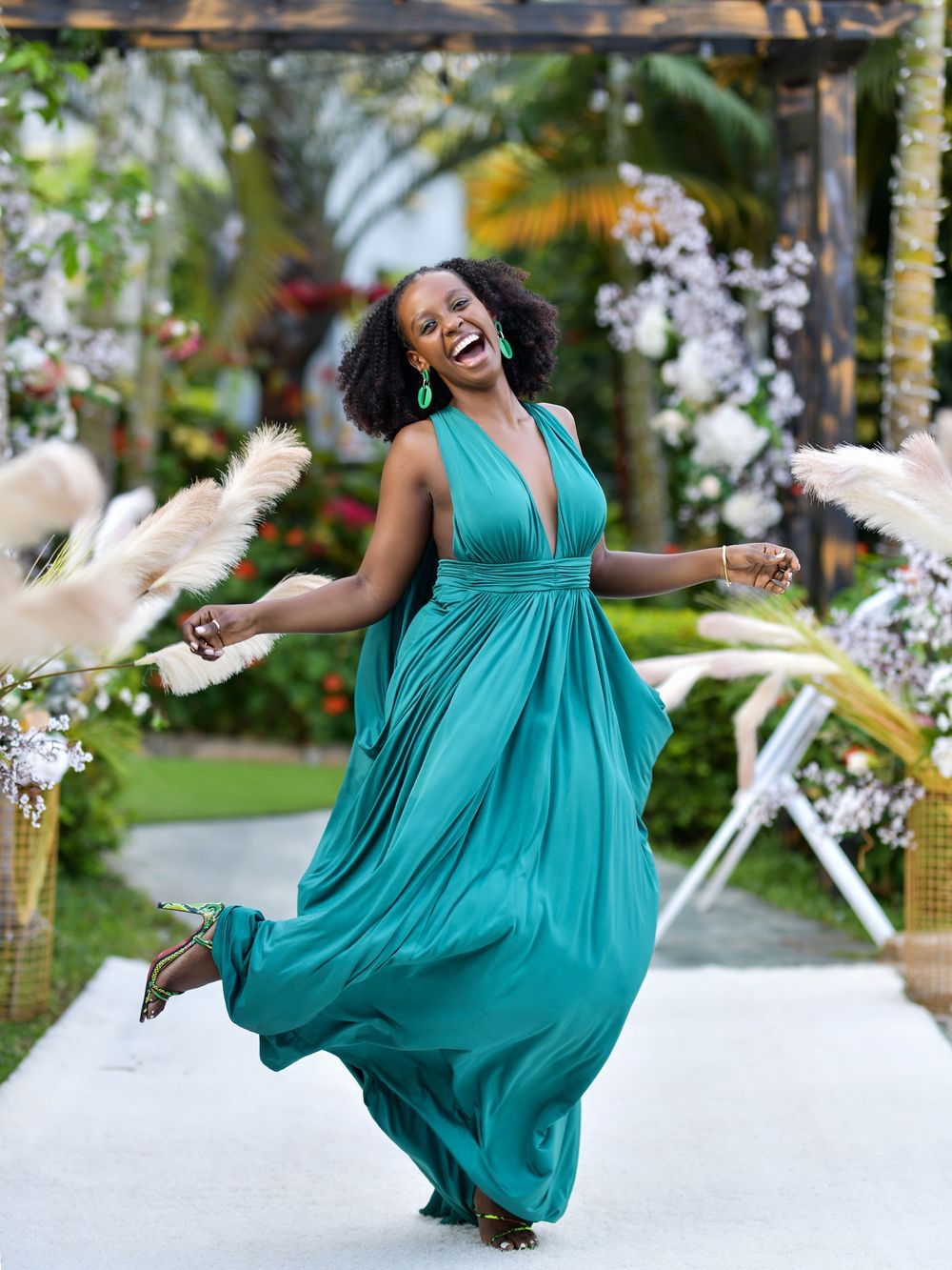 Person in flowing turquoise gown twirling joyfully in garden setting with pampas grass decorations.
