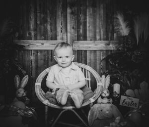 Black and white portrait of a baby sitting in a vintage chair with rustic backdrop.