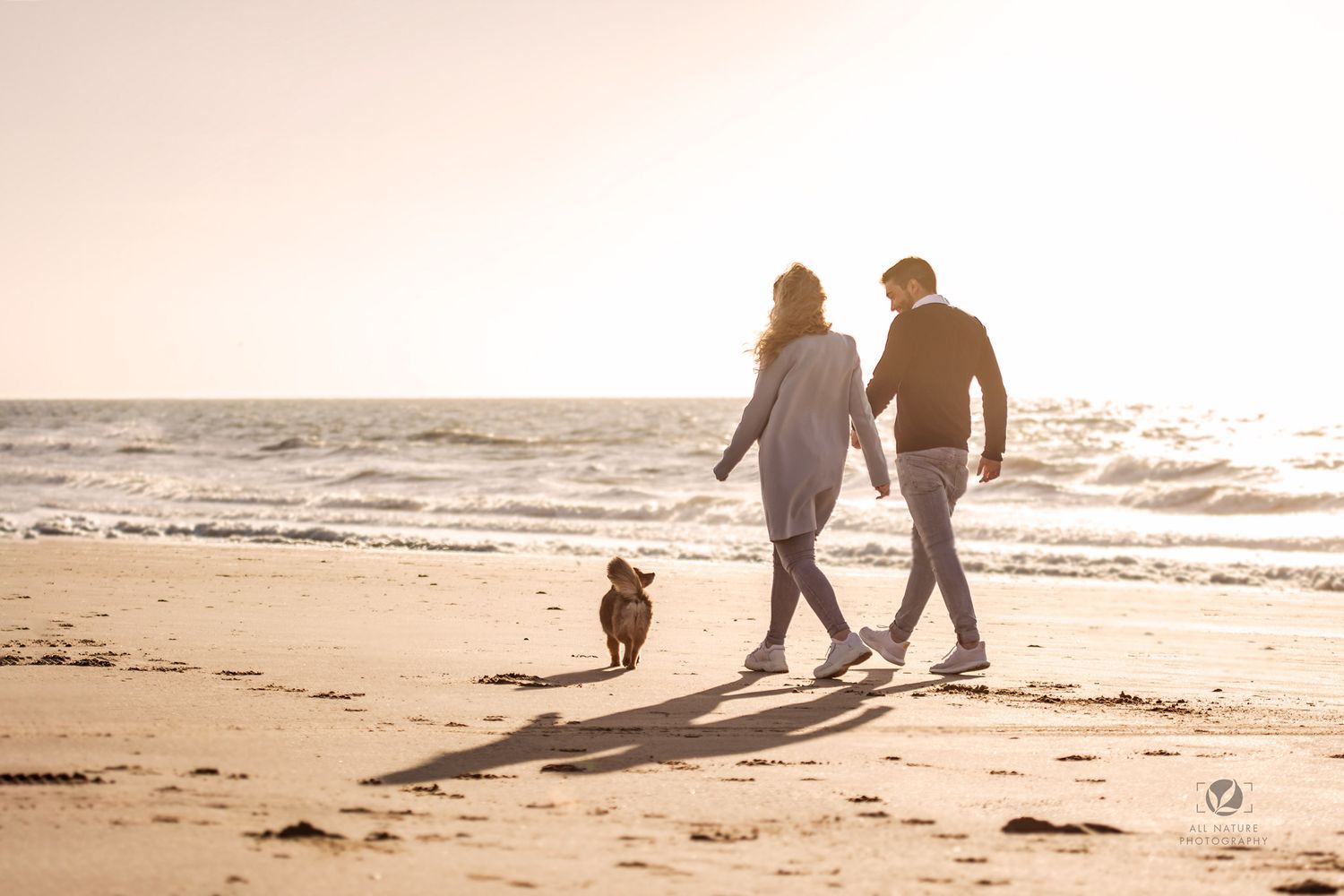 Couple walking dog on beach at sunset casting long shadows on sand.