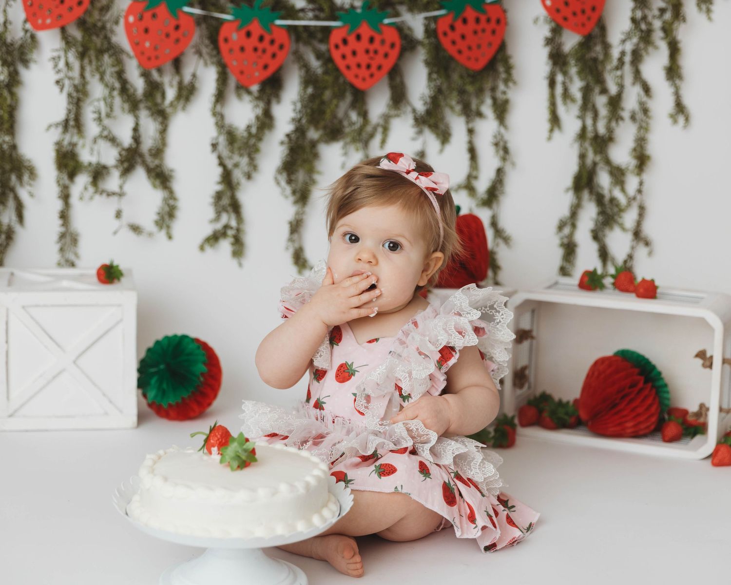 Baby in strawberry print dress with cake during first birthday photoshoot with festive decorations.