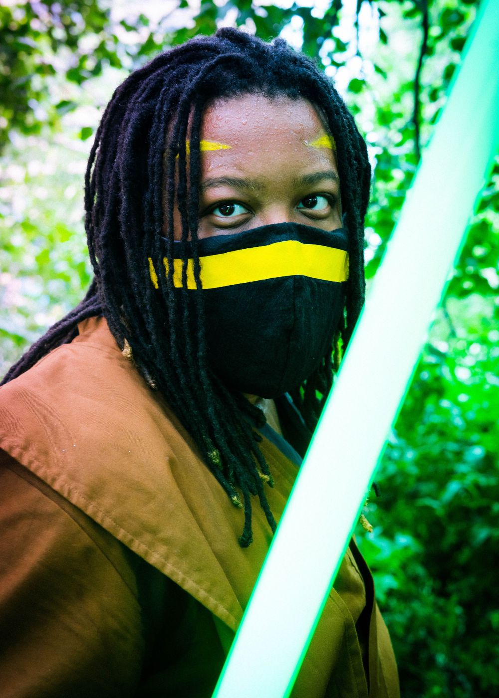Cosplayer in brown jacket with dreadlocks wearing a black and yellow mask holds a green lightsaber outdoors.