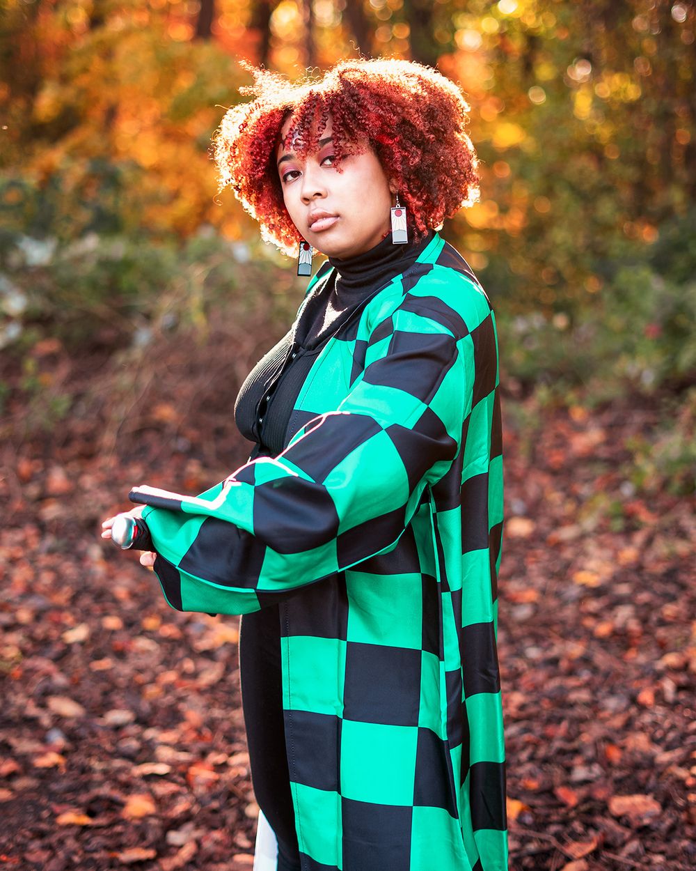Woman in green and black checkered coat with red curly hair poses in an autumn forest setting.