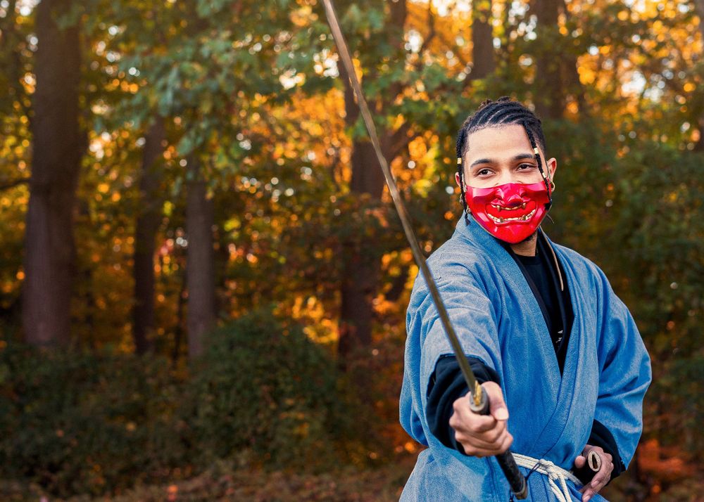 Person in blue kimono wearing red mask holds a katana sword in an outdoor forest cosplay shoot.