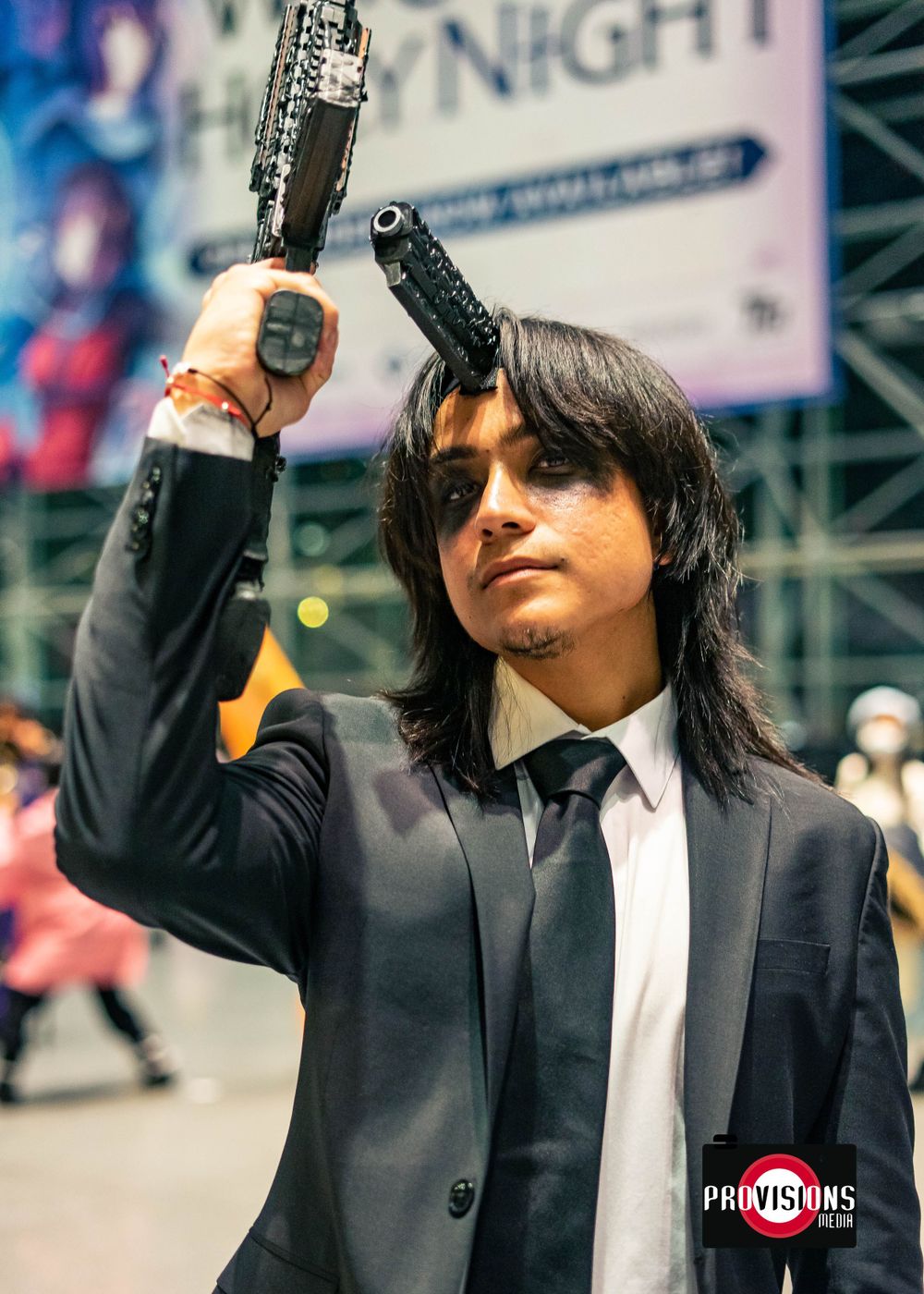A cosplayer dressed in a black suit aims two guns upward at a busy convention.