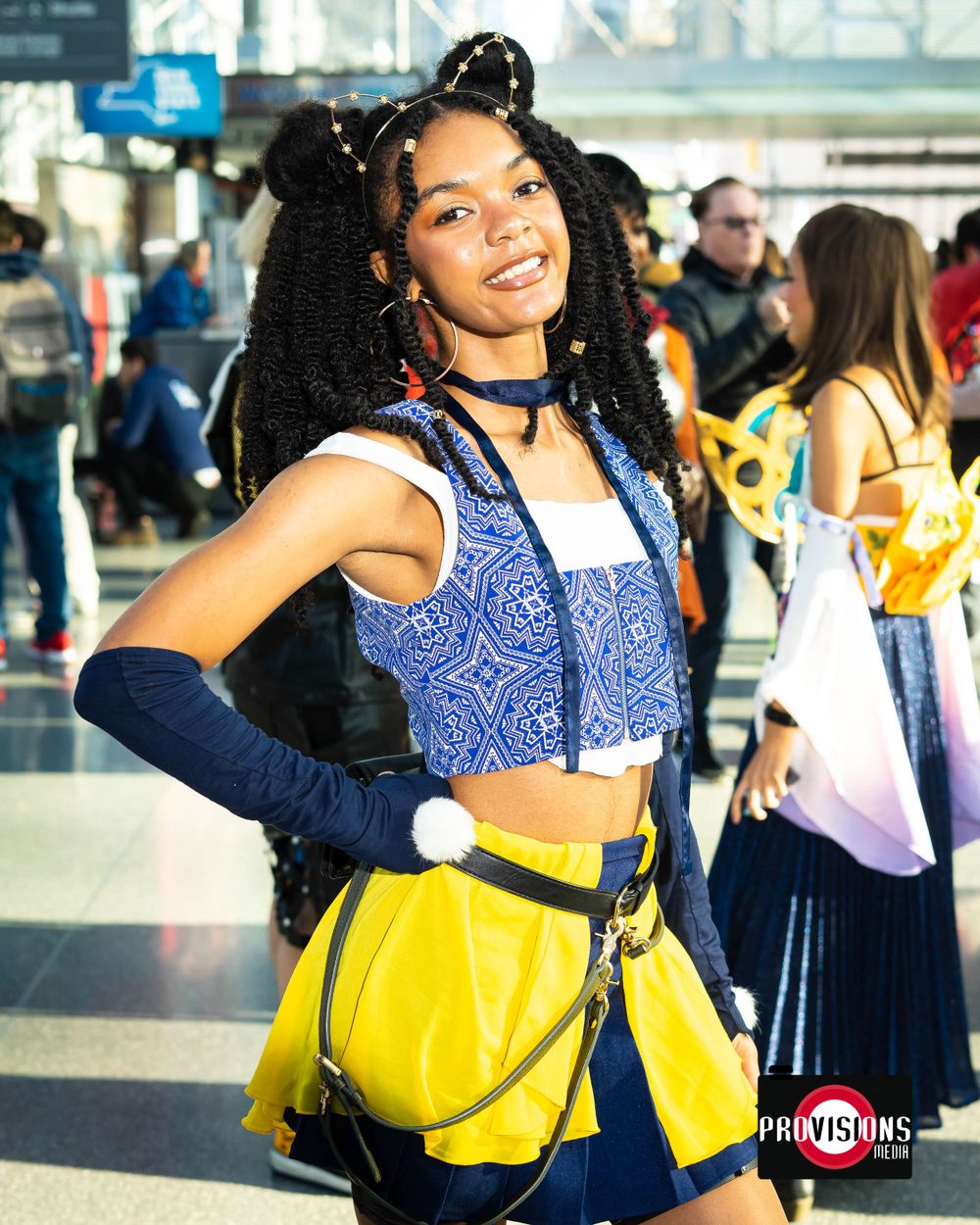 A smiling woman in a blue and yellow cosplay outfit poses at a convention center.