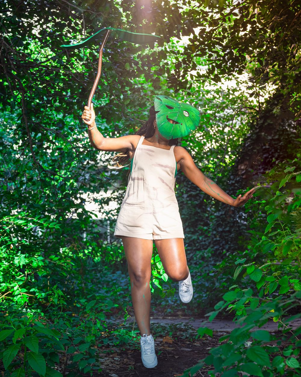 Person jumping in a forest path wearing a green leaf mask and holding a branch in lush greenery.