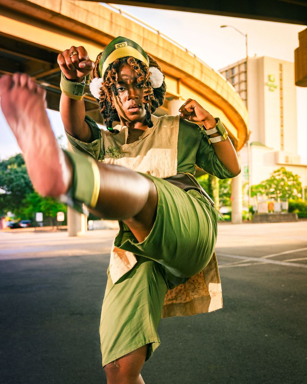 Person cosplaying as Toph from Avatar in green outfit performing a high kick under an urban overpass.