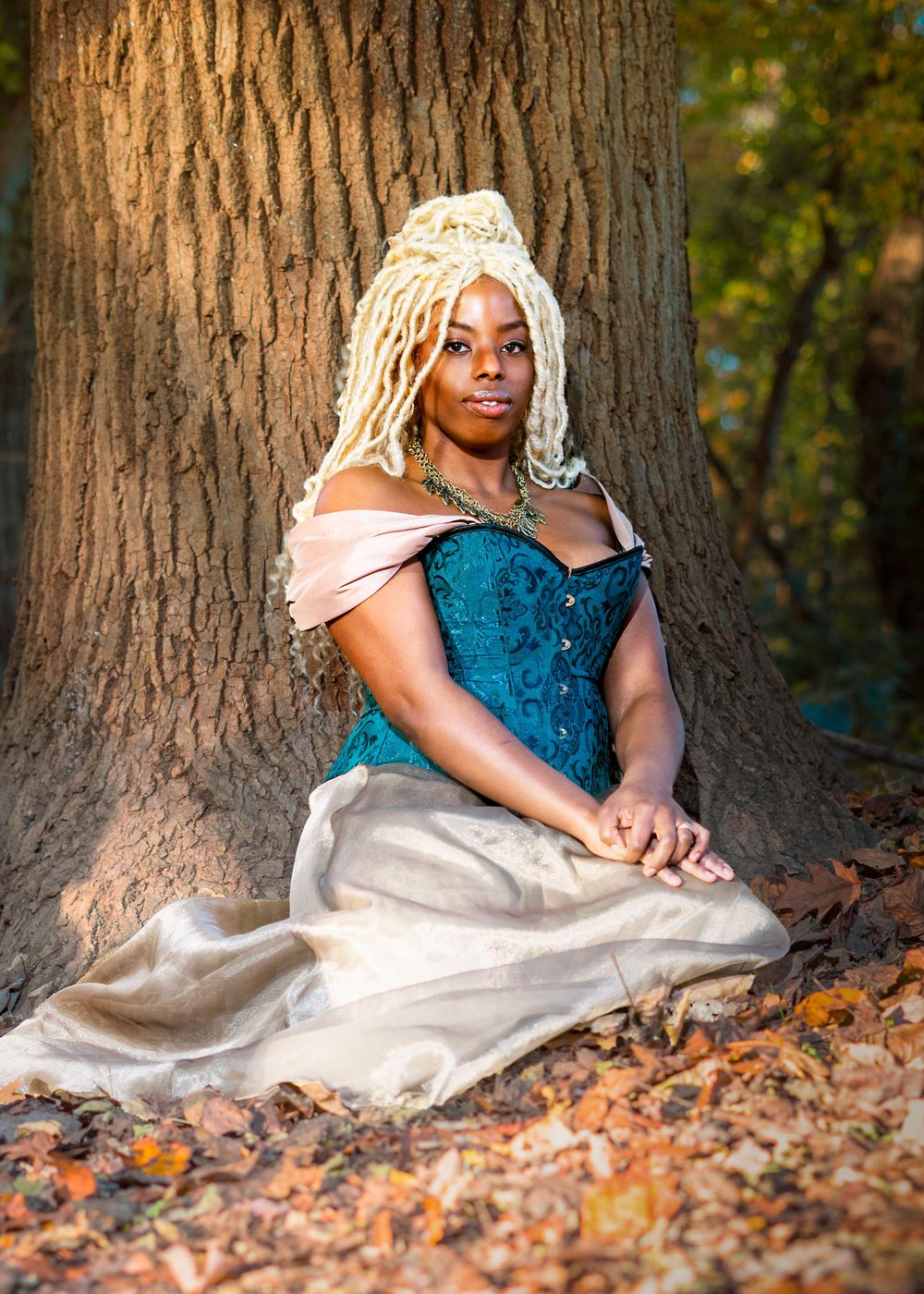 Woman in teal corset gown with blonde locs sits regally against a large tree in autumn woods.