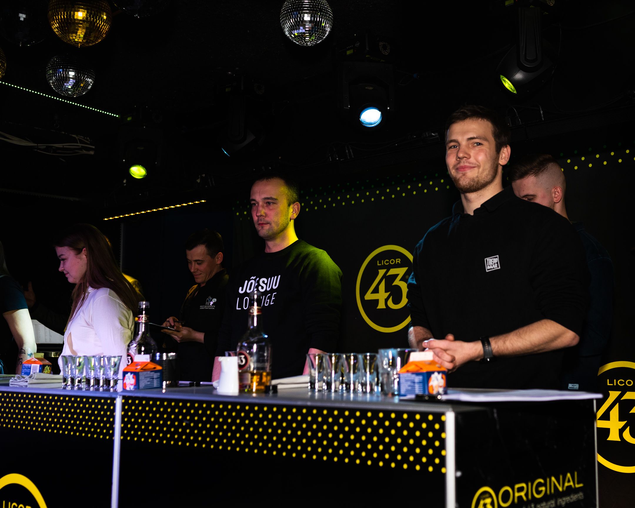 Staff working behind an illuminated bar counter with Licor 43 branding and disco balls overhead.