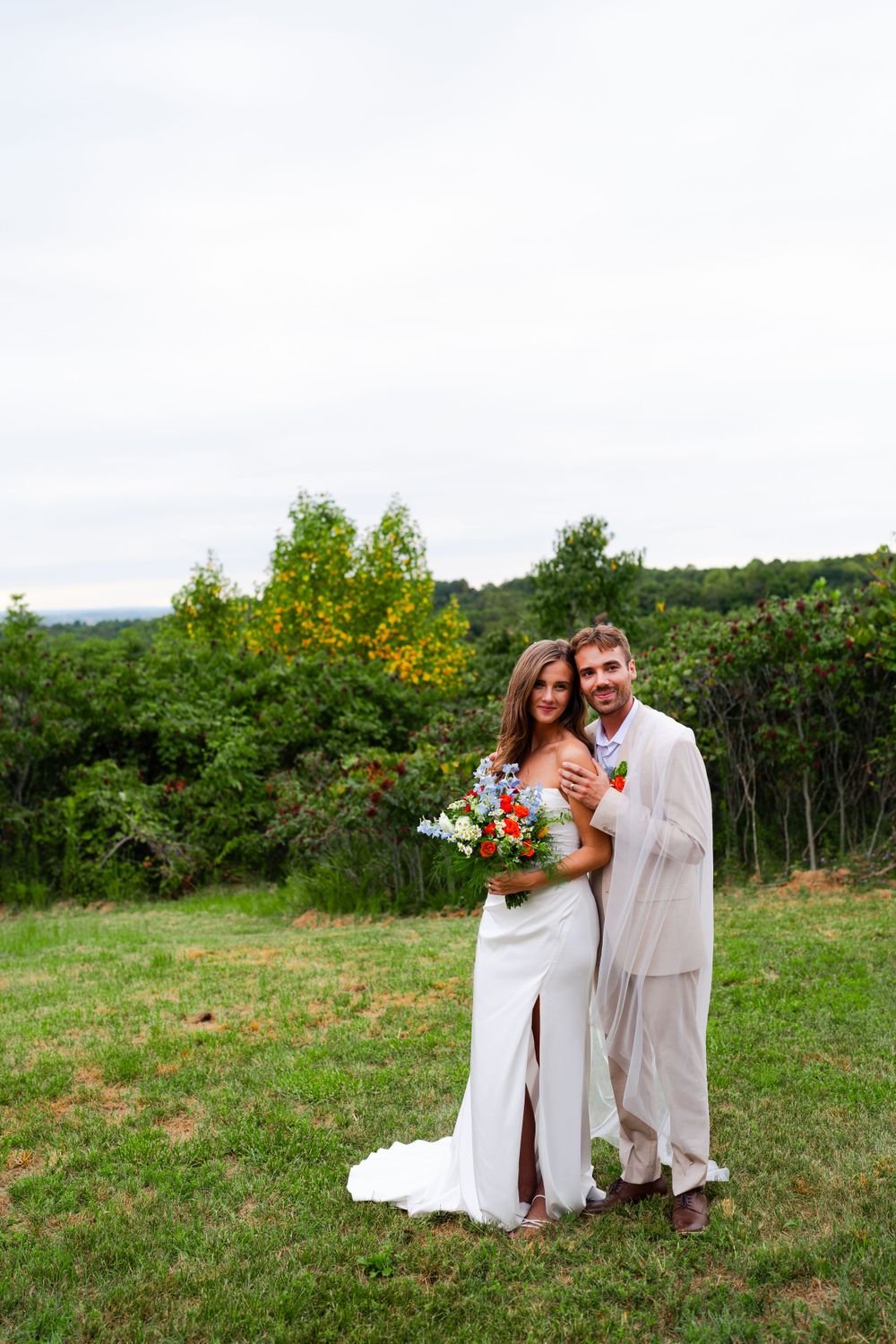 A couple poses for wedding photos in a scenic hilltop meadow setting.