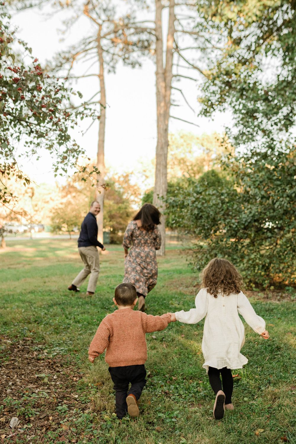 Two children running together on a grassy path surrounded by pine trees at sunset.