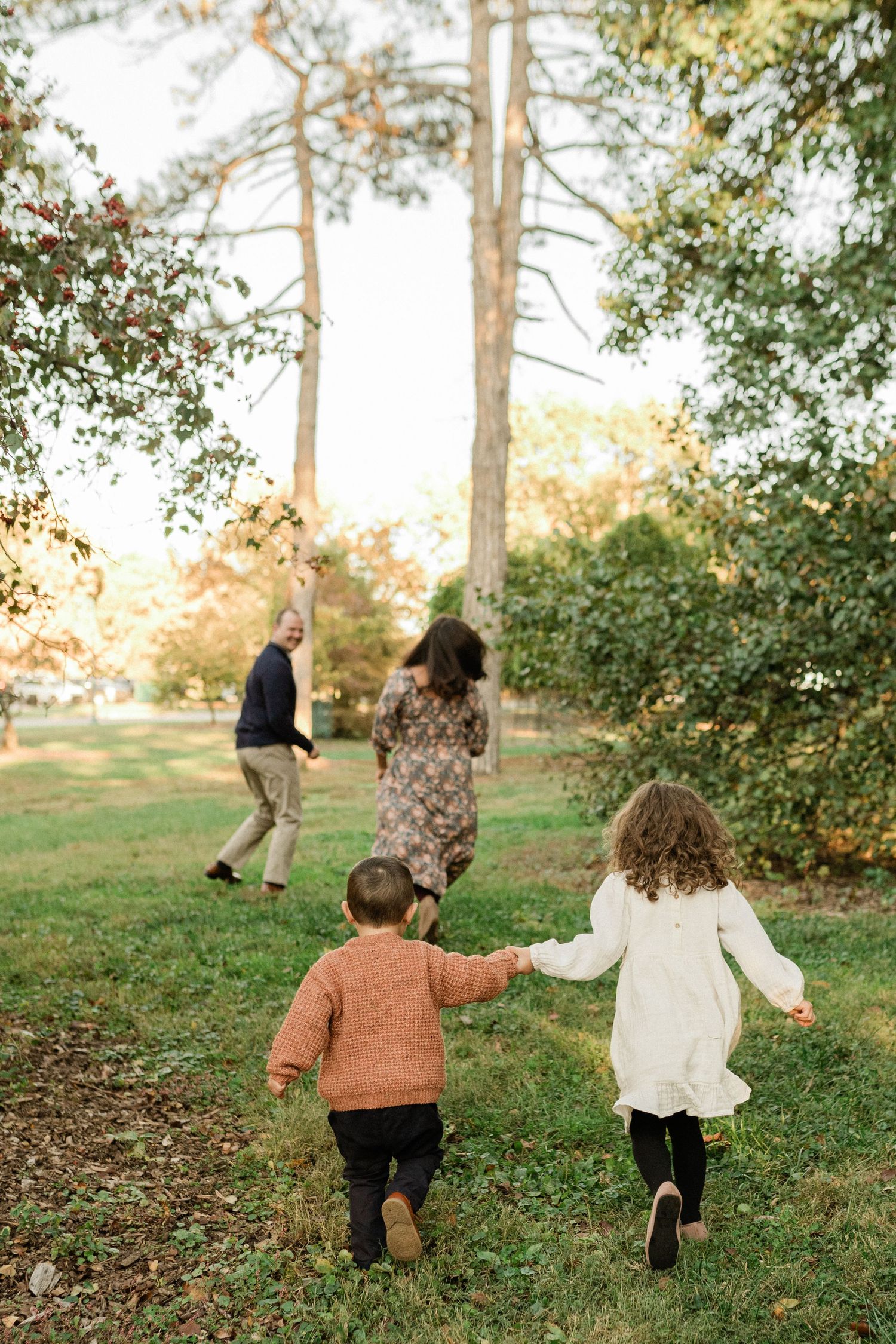 Two children running together on a grassy path surrounded by pine trees at sunset.