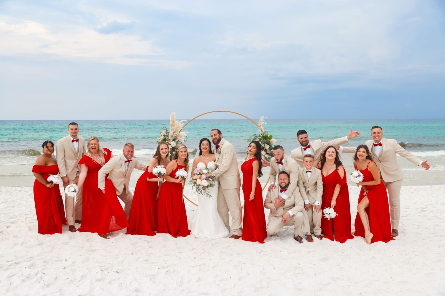 Wedding party poses on beach with bridesmaids in red dresses and groomsmen in beige suits.