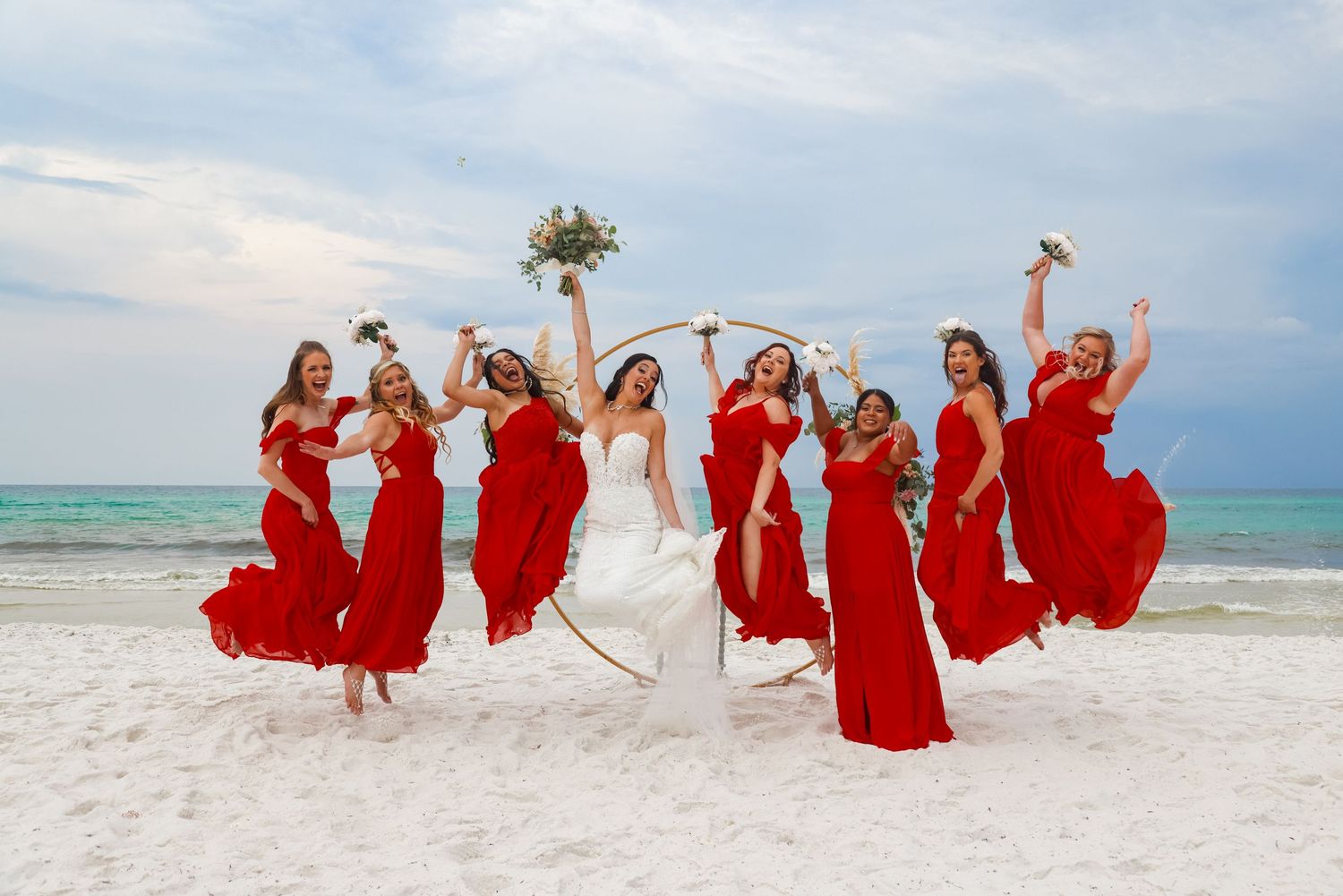 Bridal party jumps joyfully on beach with bridesmaids wearing flowing red dresses.