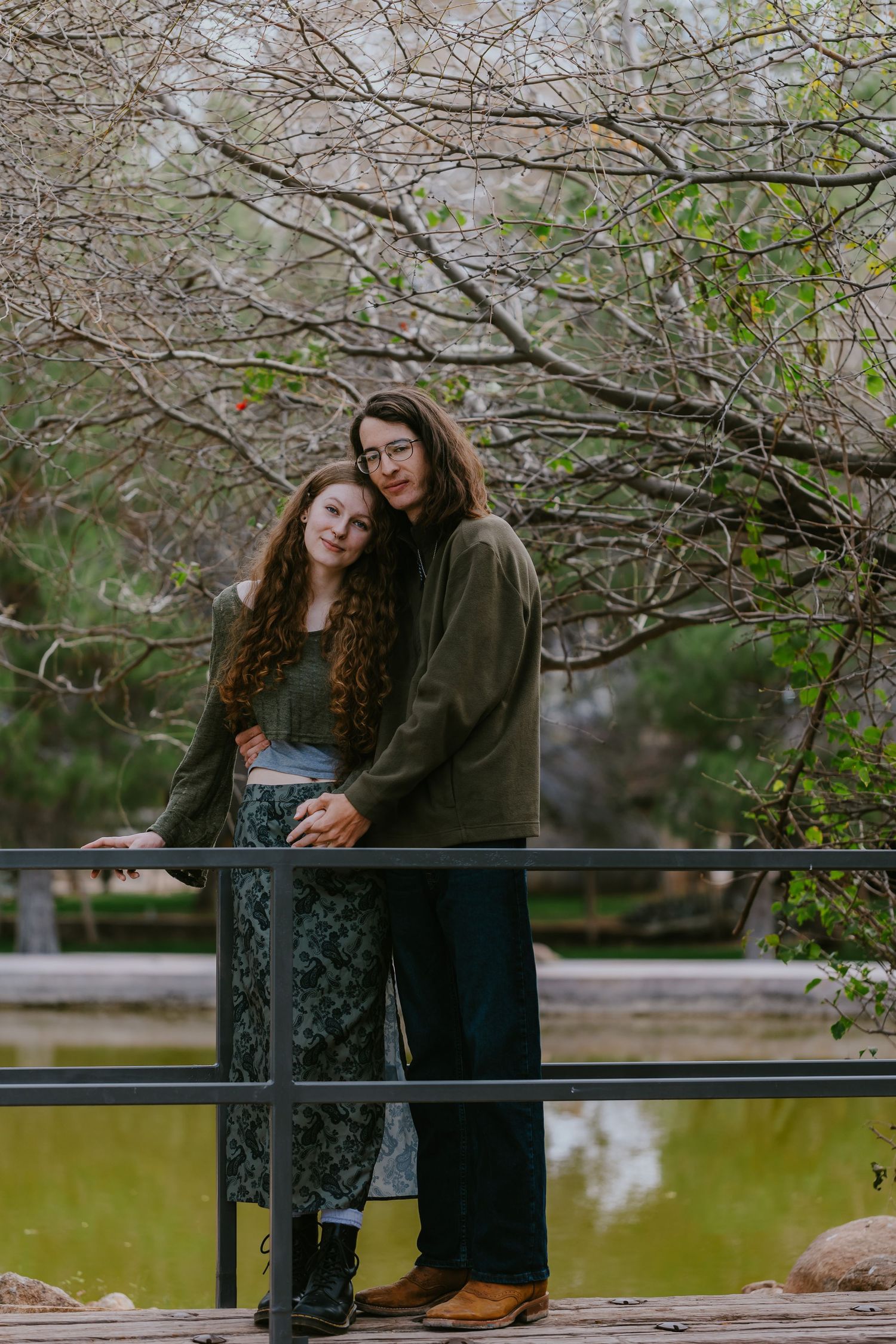 A couple sits together on a railing overlooking a pond surrounded by spring trees.