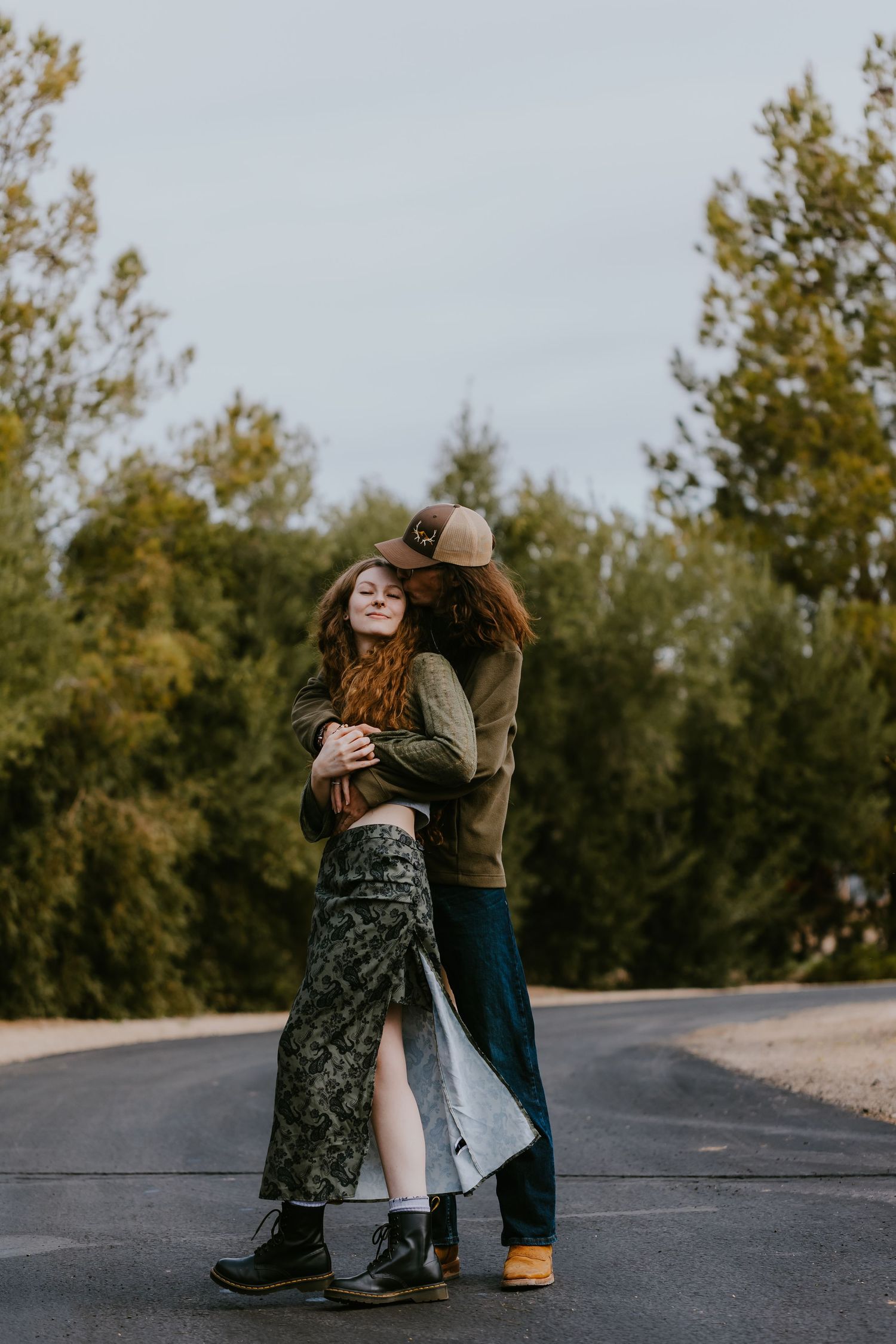 A romantic moment captured on a rural road with tall trees in the background.