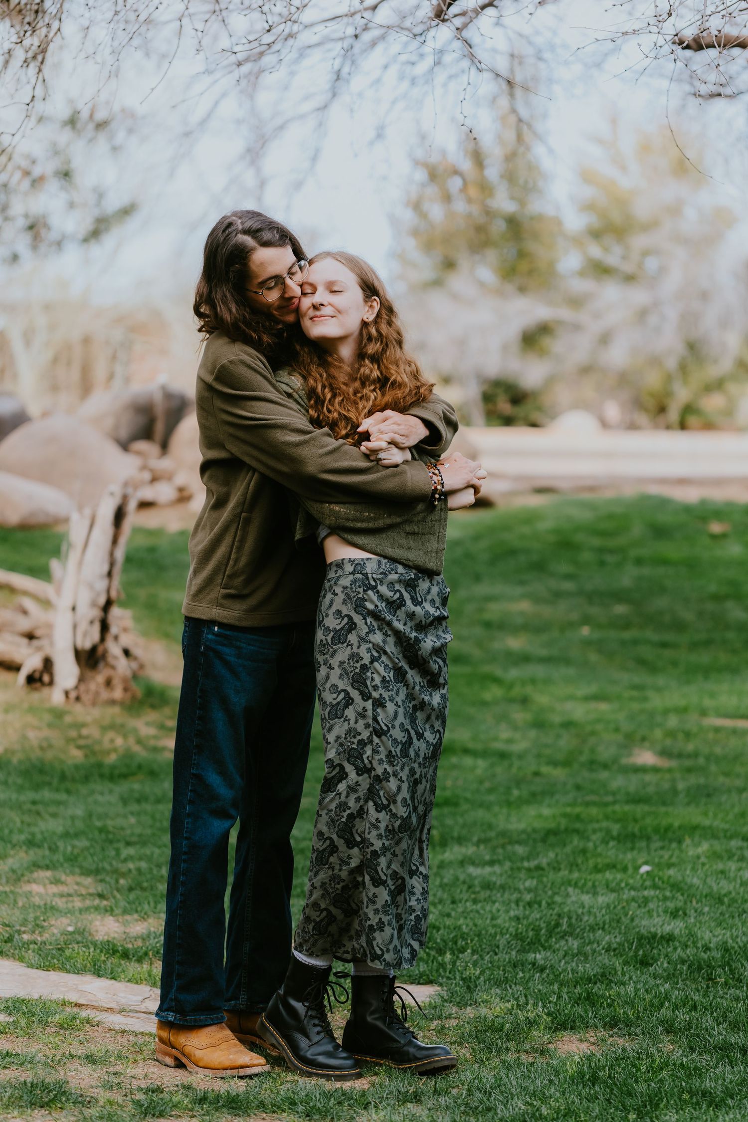 Couple embracing on green lawn with bare trees in background.