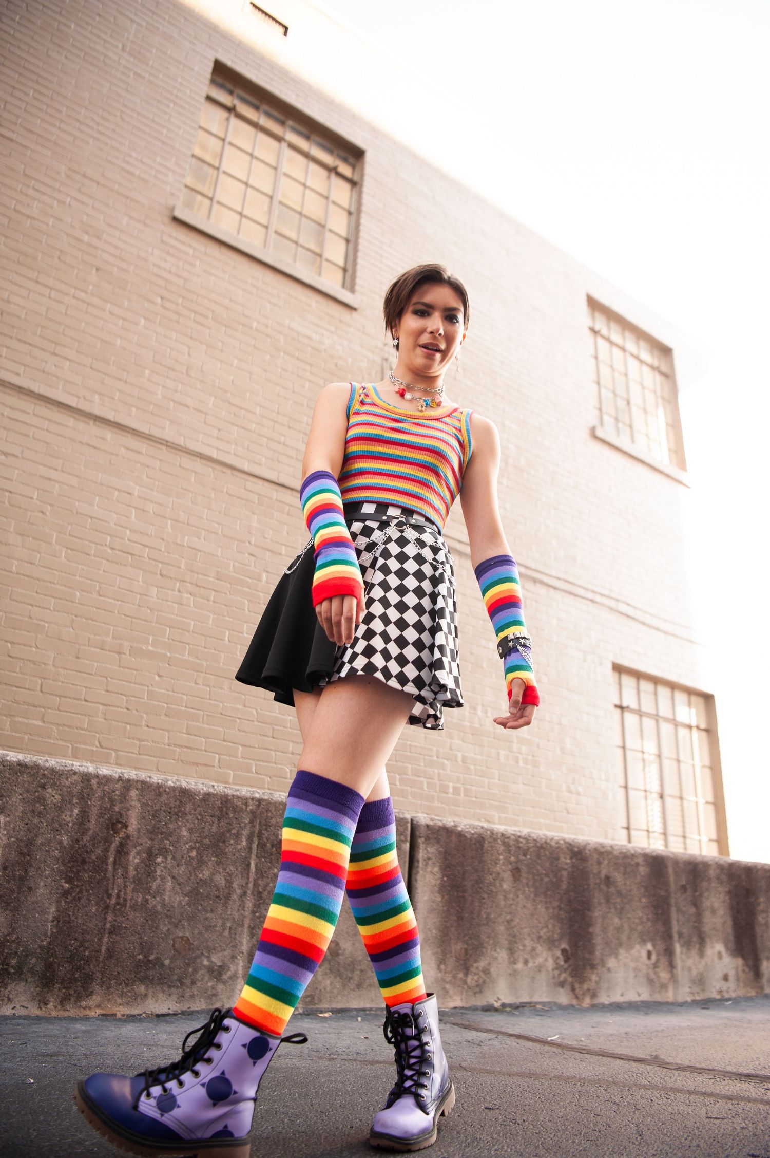 A street fashion look featuring rainbow socks, checkerboard skirt, and purple boots against an urban backdrop.
