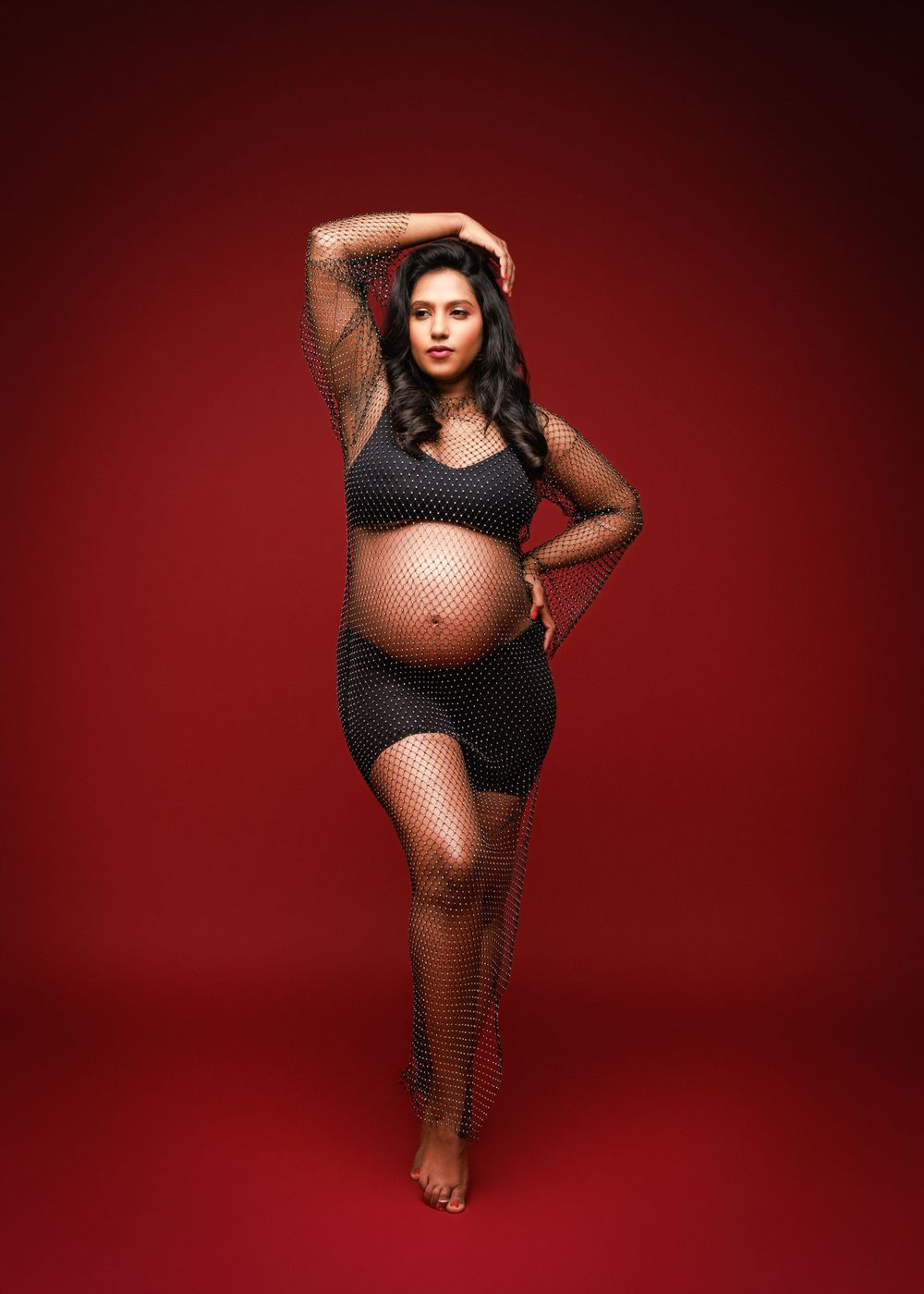 Artistic maternity pose in black mesh outfit against red backdrop.