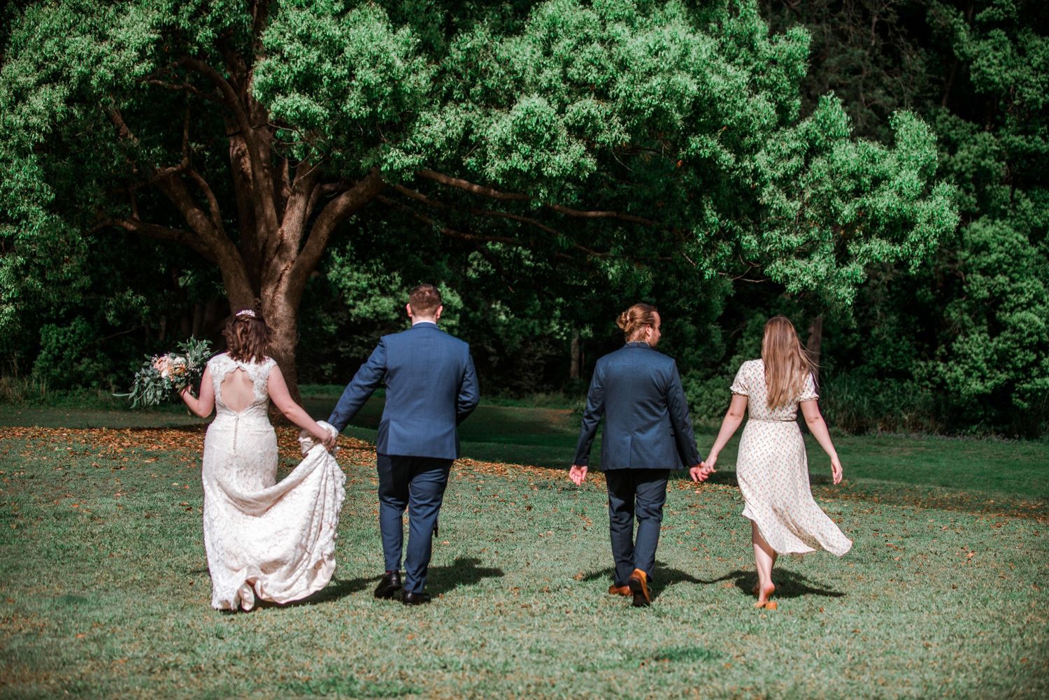 Four wedding guests walk hand in hand across a grassy lawn.
