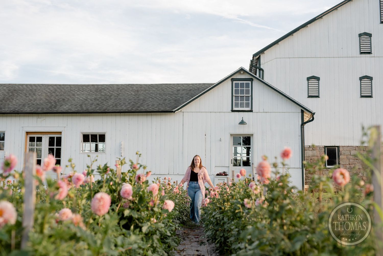Person walking through a flower garden path with white barn buildings in the background.
