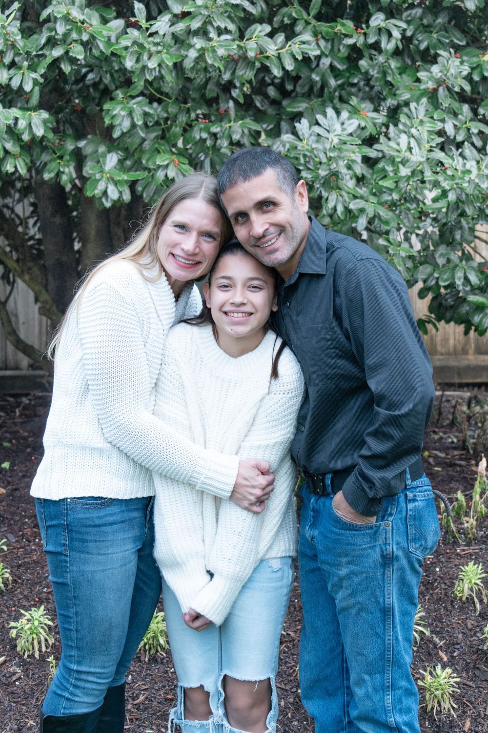 A family wearing white and blue sweaters poses together against green foliage in an outdoor portrait.