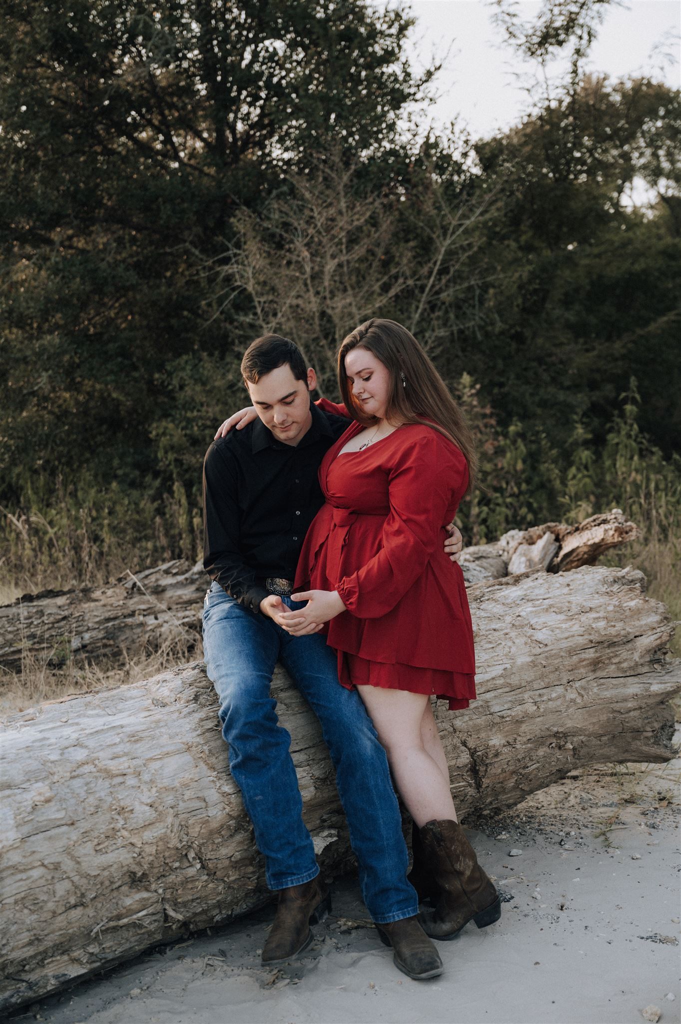 Couple sits together on a fallen log while looking at something on a device.