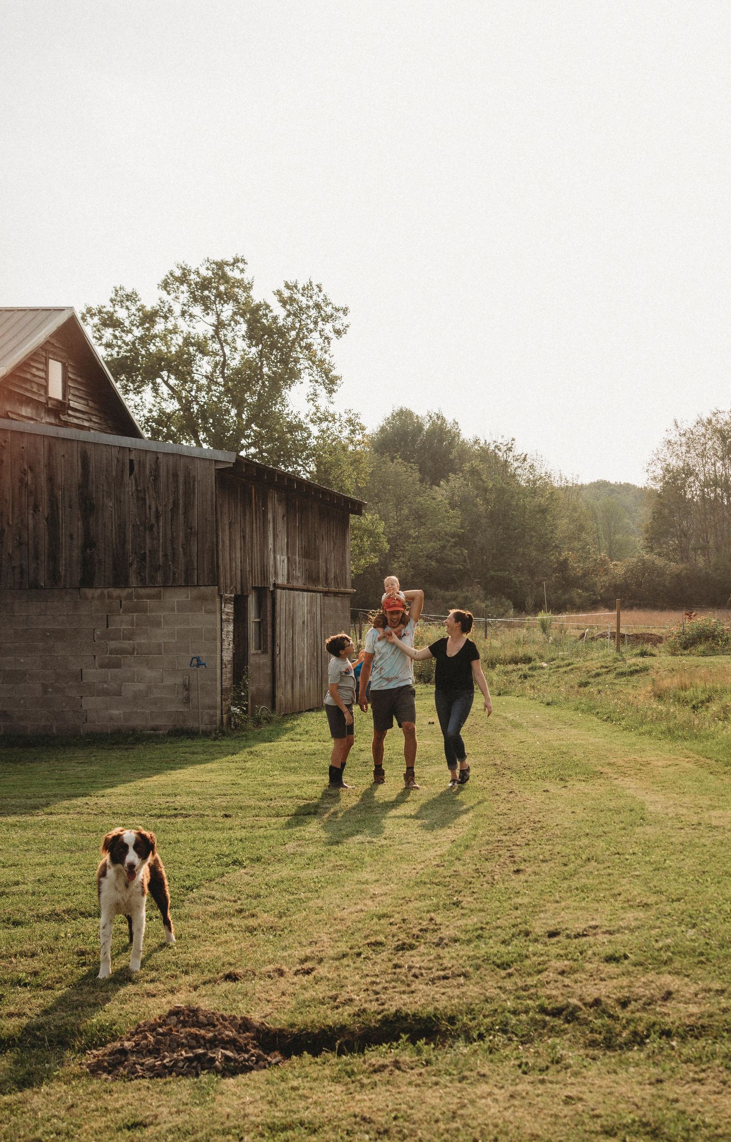 People walking together on a grassy path near an old barn with a dog following behind.