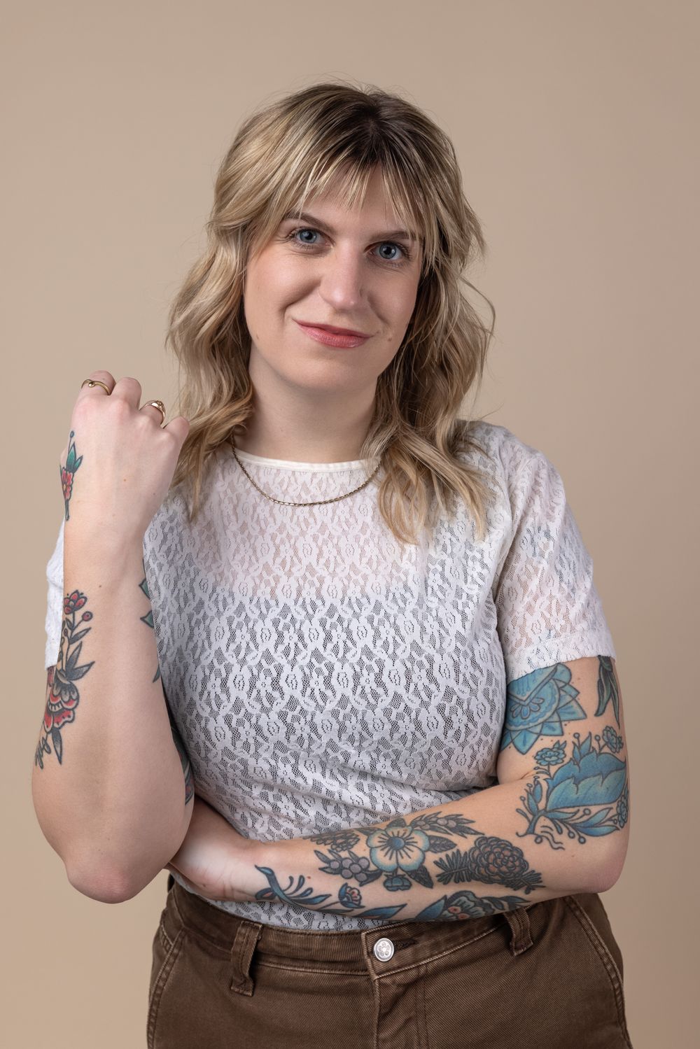 Blonde woman with colorful tattoos on both arms wearing white lace top and brown pants posing against beige background.