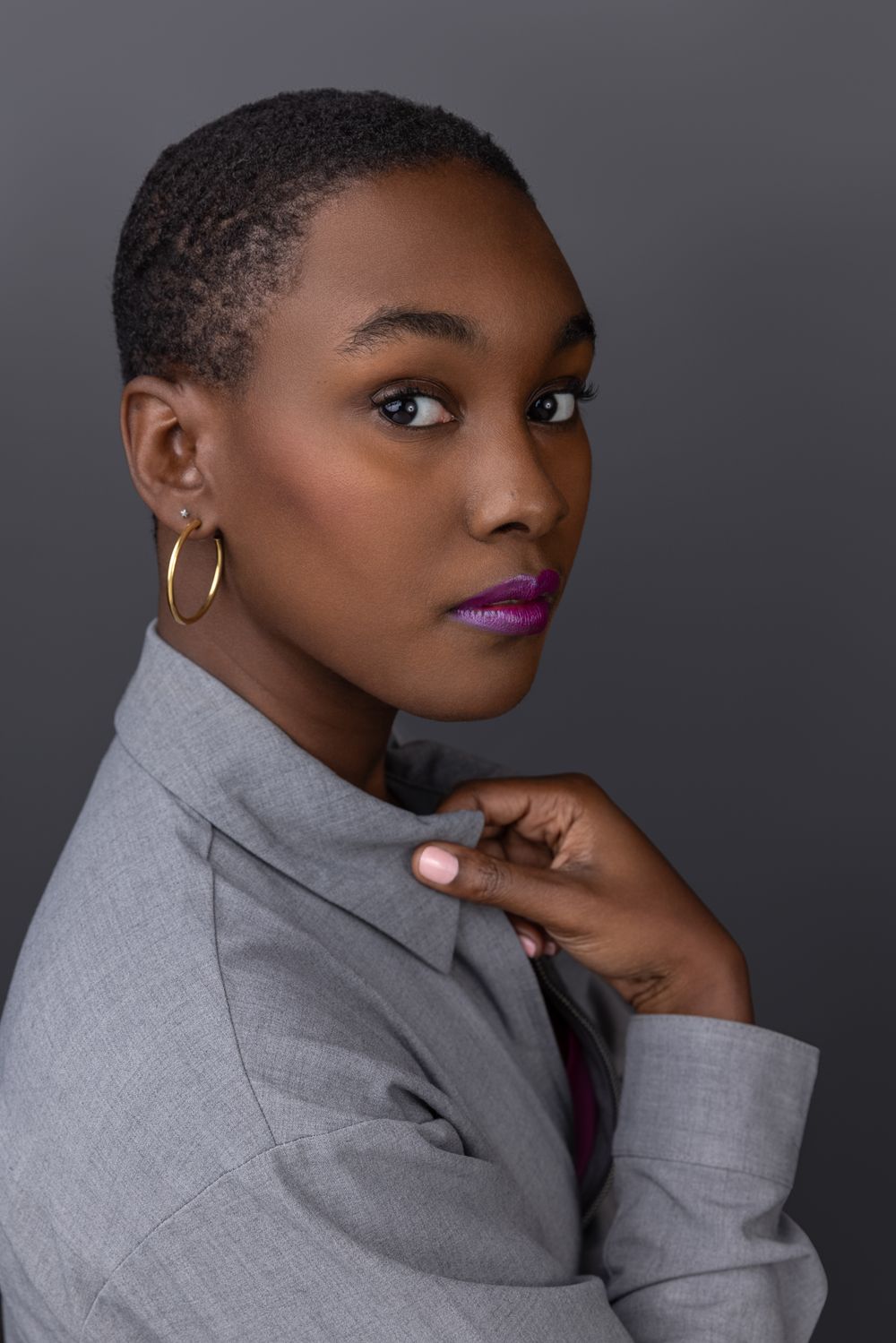 Stylish Black woman with short hair wearing gray blazer and hoop earrings posing against gray background.