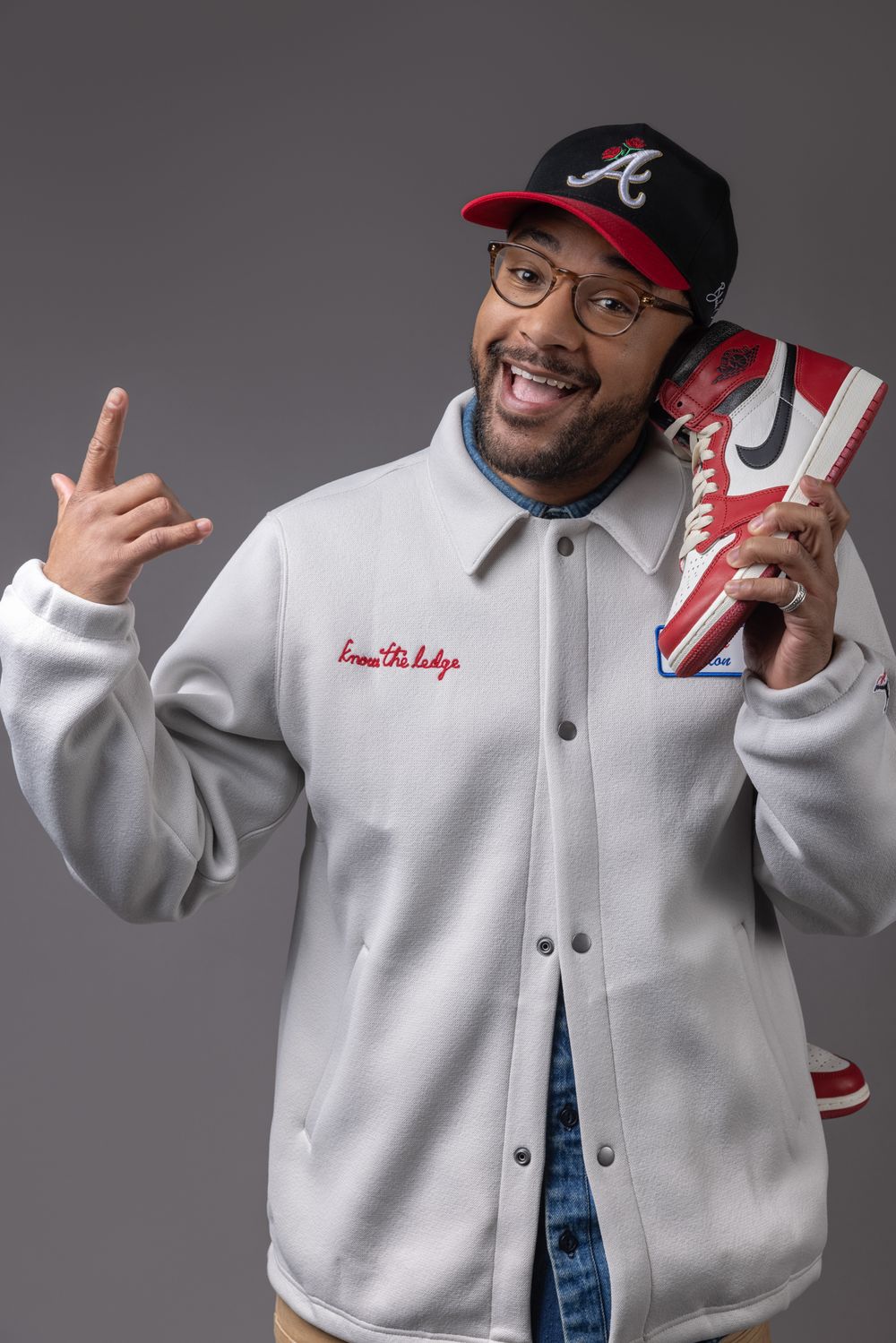 Cheerful man in white jacket holds sneaker over shoulder, wearing Atlanta Braves cap and glasses.