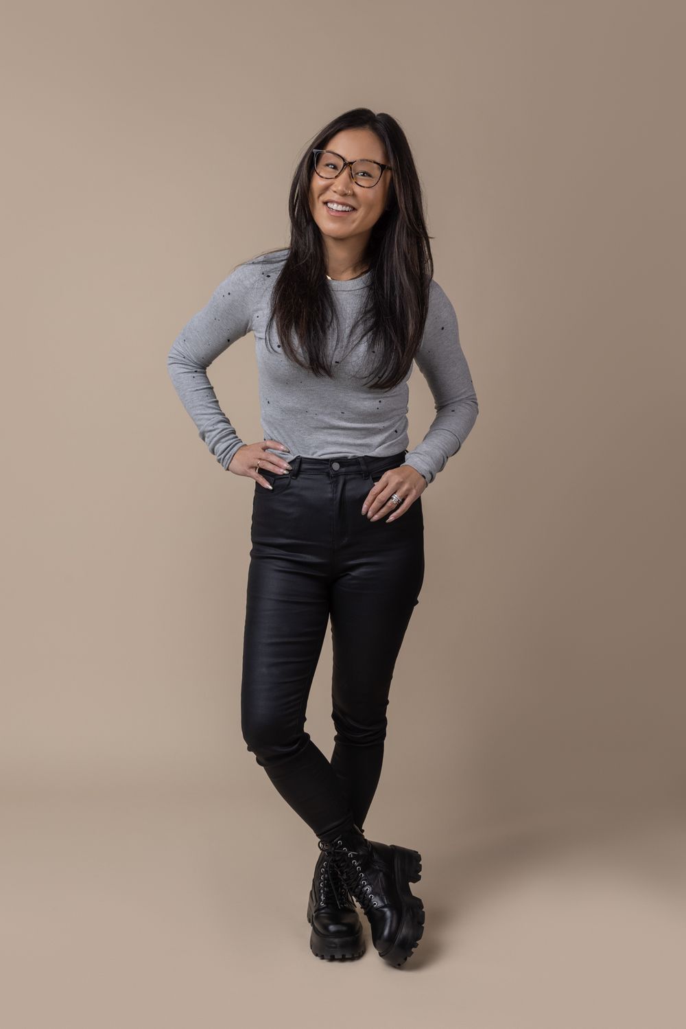Smiling Asian woman in gray top and black jeans stands confidently wearing glasses and boots.