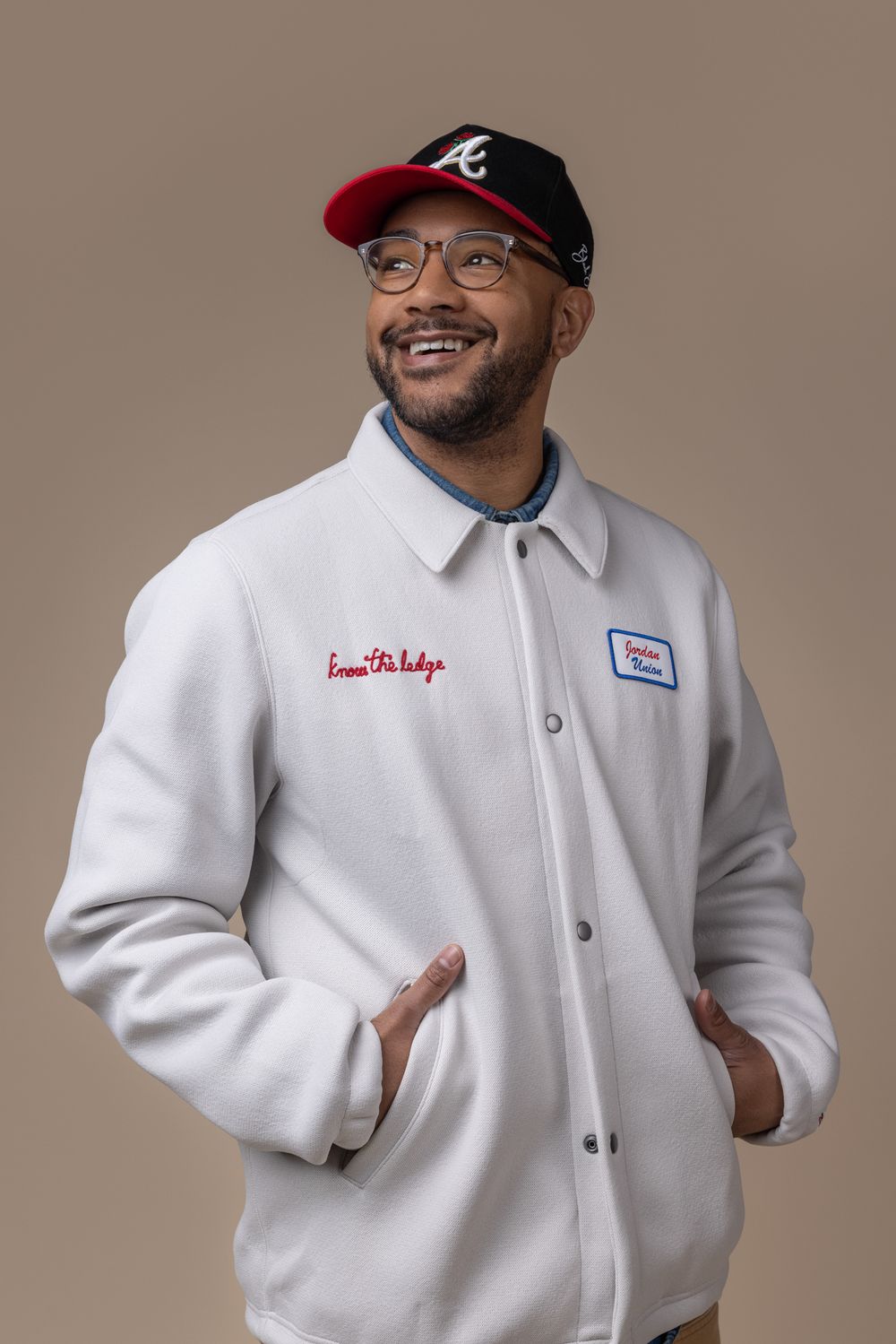 Smiling man in white varsity jacket and Atlanta Braves cap poses with hands in pockets.