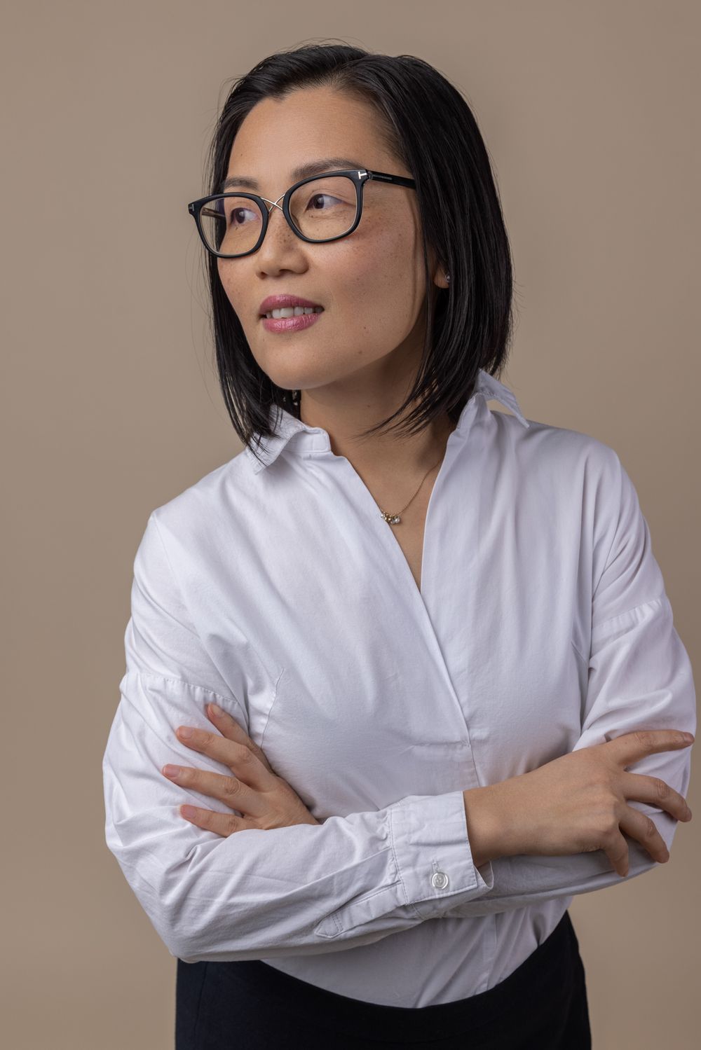 Asian woman with black-rimmed glasses in white button-up shirt standing with arms crossed against beige background.