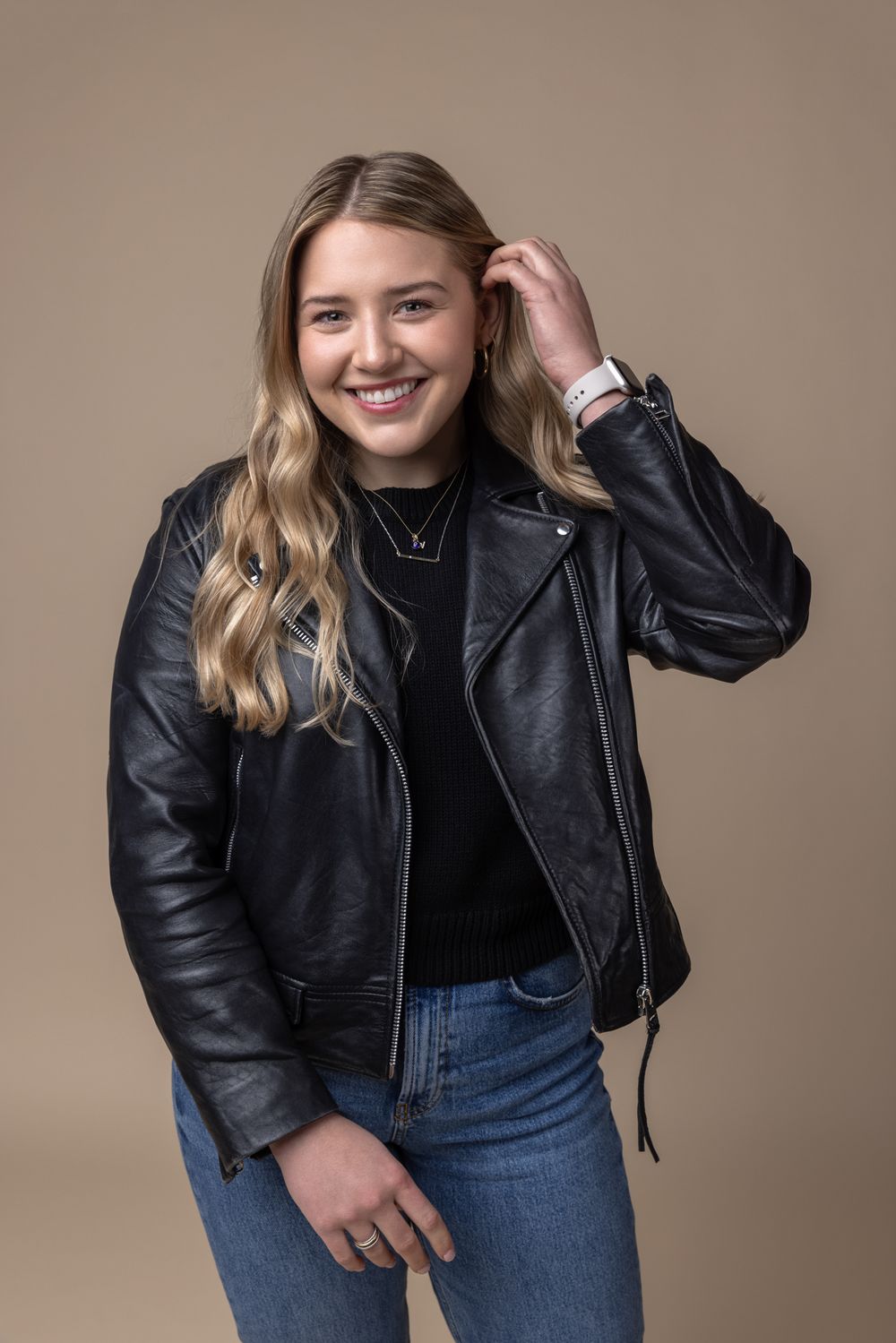 Smiling blonde woman in black leather jacket and jeans touching her hair against beige background.
