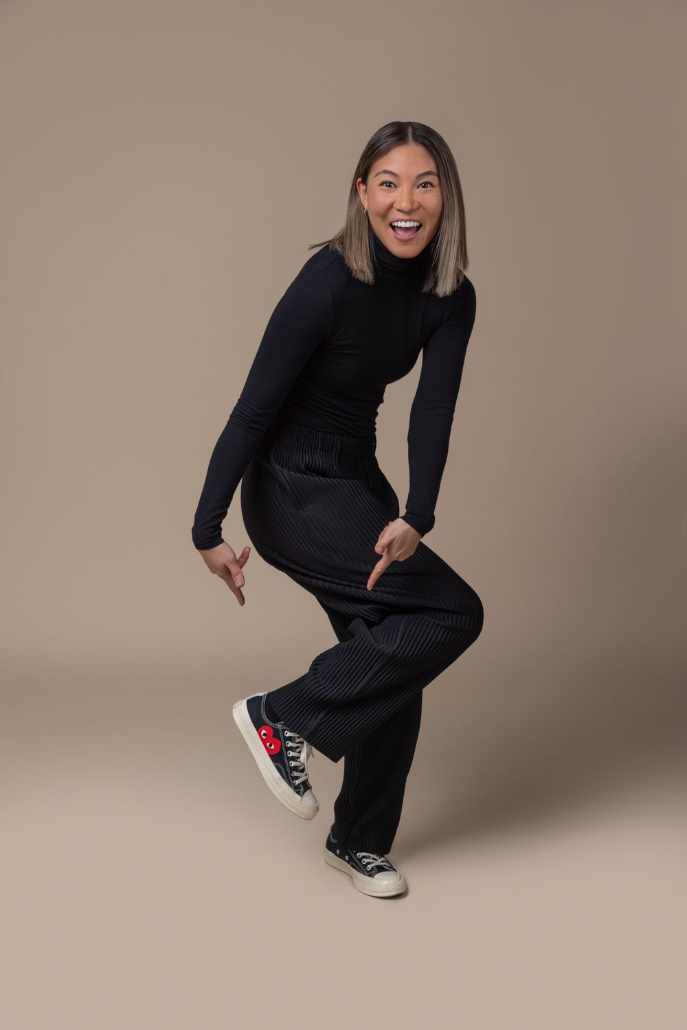 Joyful Asian woman in black turtleneck and wide-leg pants poses dynamically in studio.
