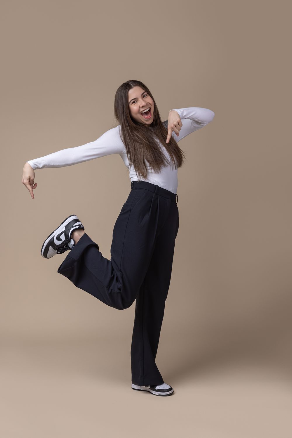 Energetic young woman in white top and black wide-leg pants dancing against beige background.
