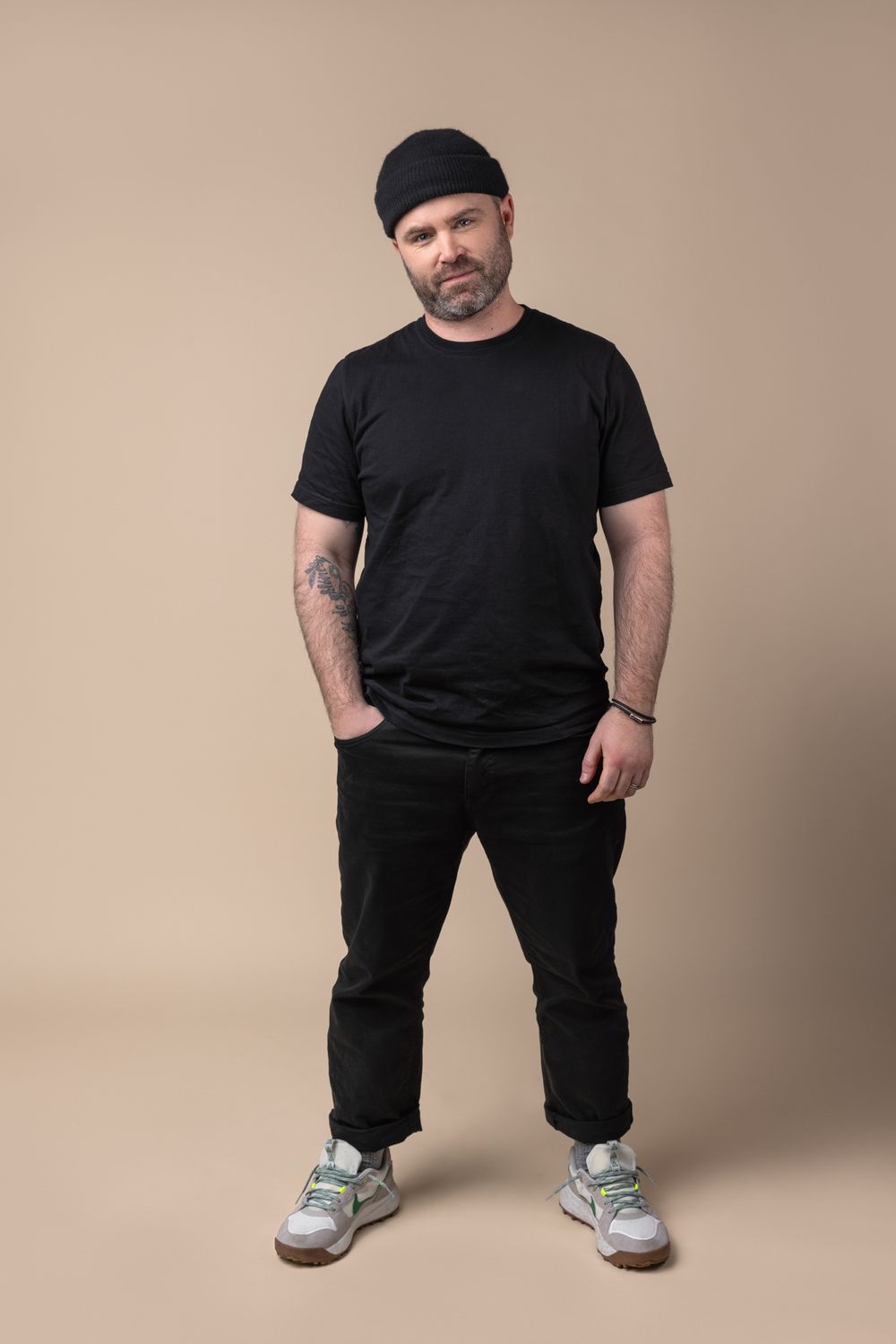 Middle-aged man in all-black outfit with beanie hat and tattoos standing with hands in pockets against beige background.