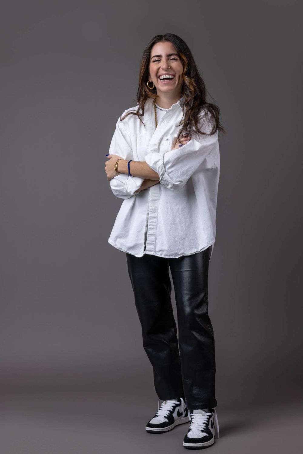 Laughing woman in white oversized shirt and black leather pants with sneakers on gray background.