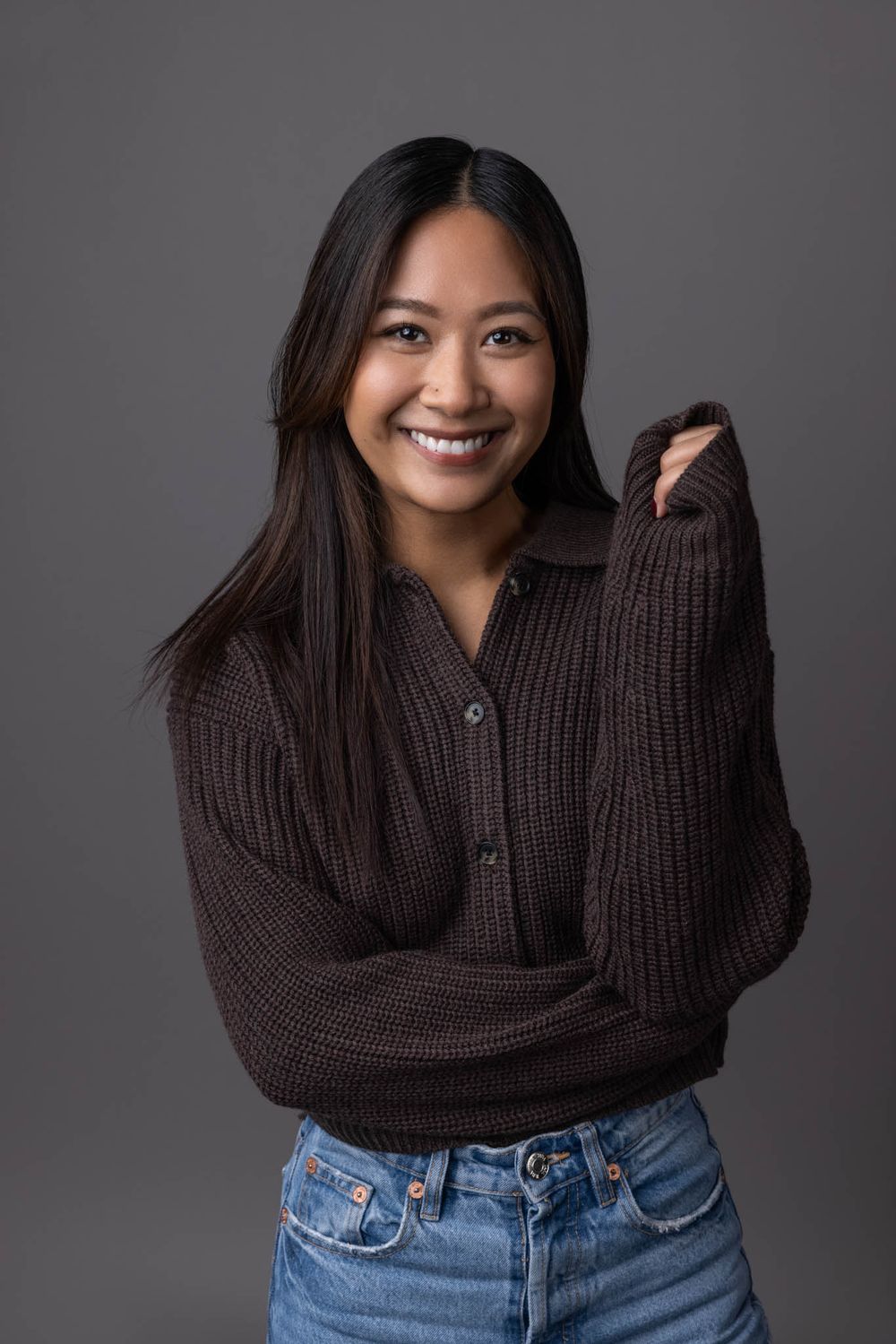 Smiling young Asian woman in dark brown knit sweater and jeans on gray background.