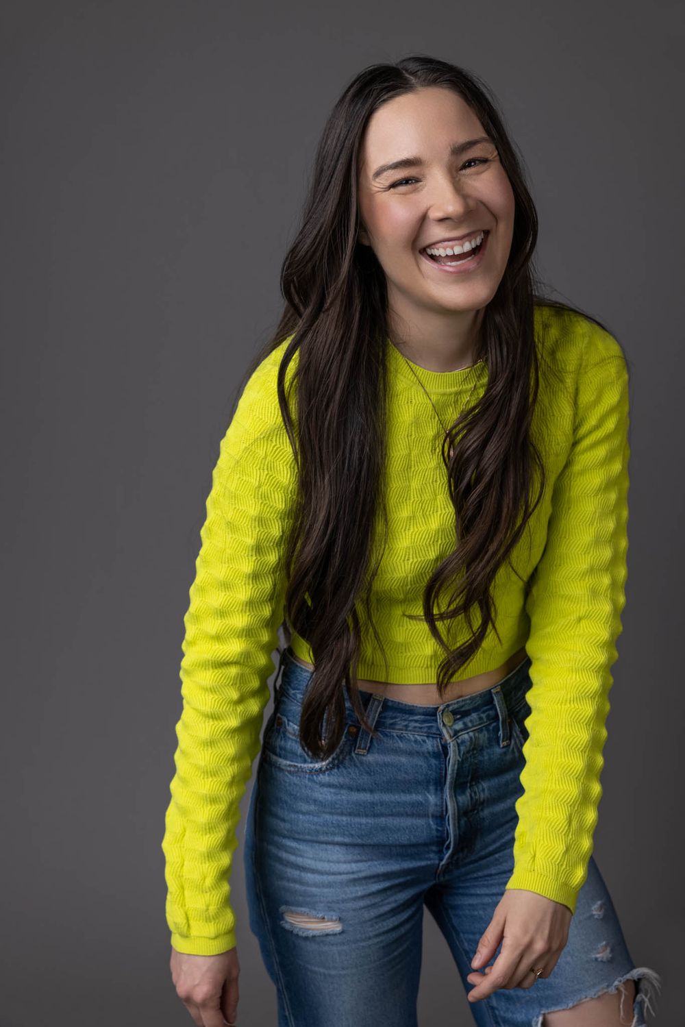 Laughing young woman in neon yellow crop sweater and denim shorts on gray background.