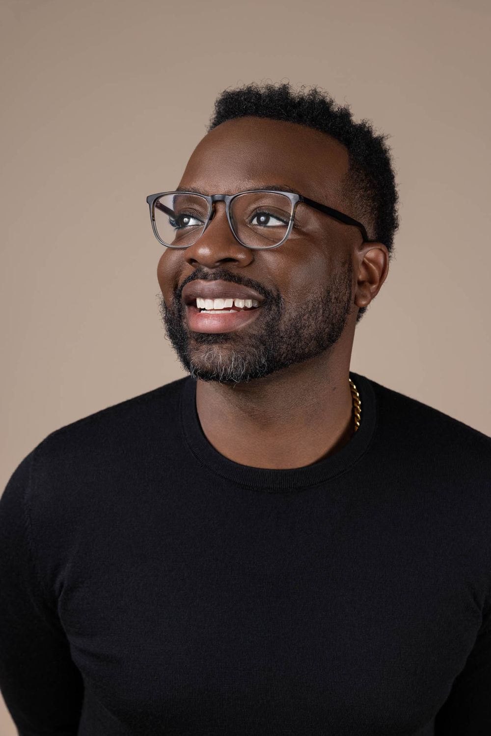 Smiling Black man with glasses and gold chain necklace wearing black sweater against beige background.