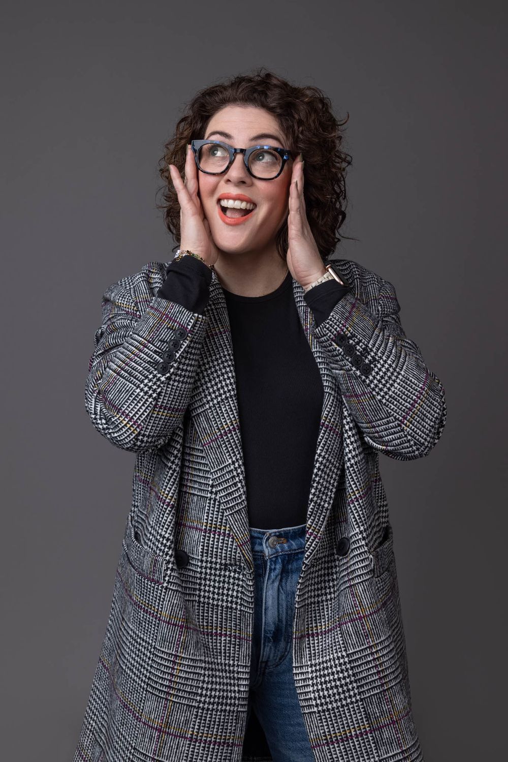 Excited curly-haired woman in plaid blazer and glasses holding face with hands against gray background.